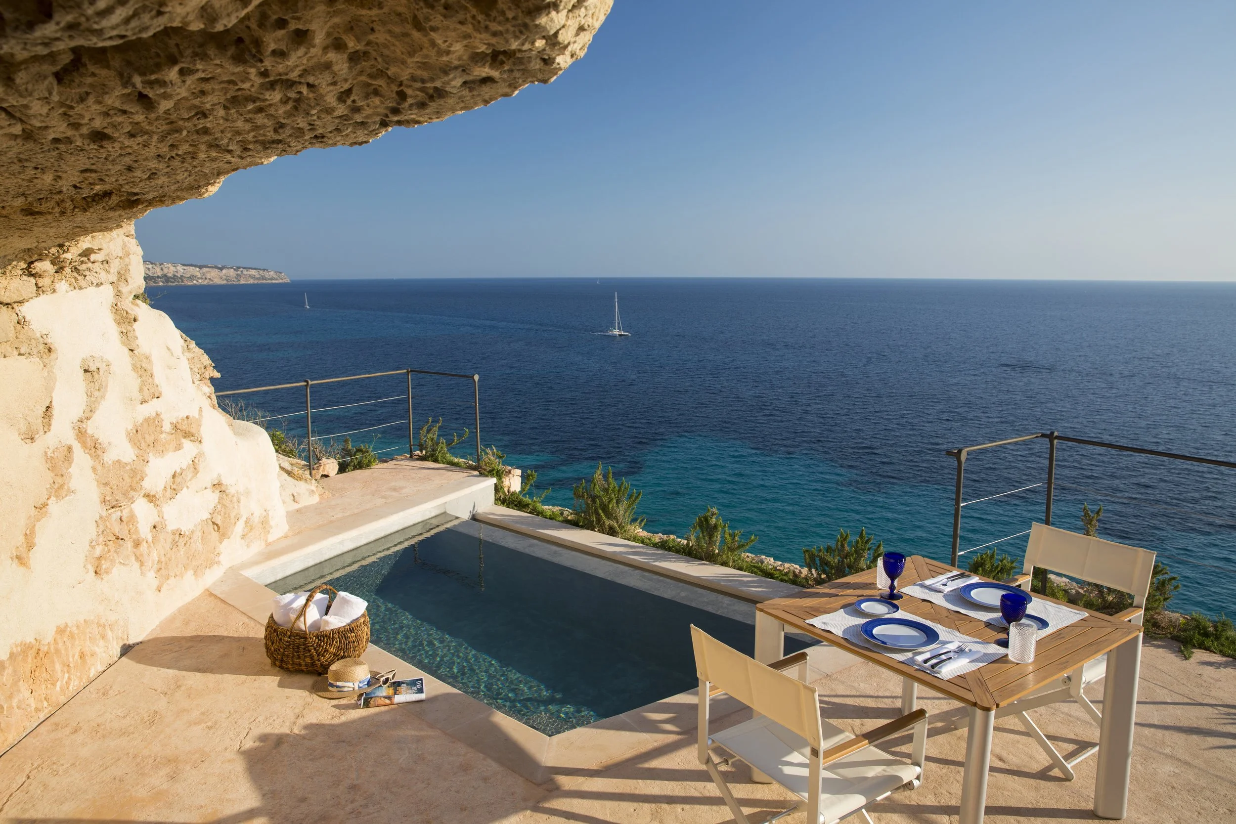 A seaside outdoor dining area with a table set for two, overlooking the ocean with sailboats. There is a small pool, a basket with towels, and a rocky overhang providing shade.