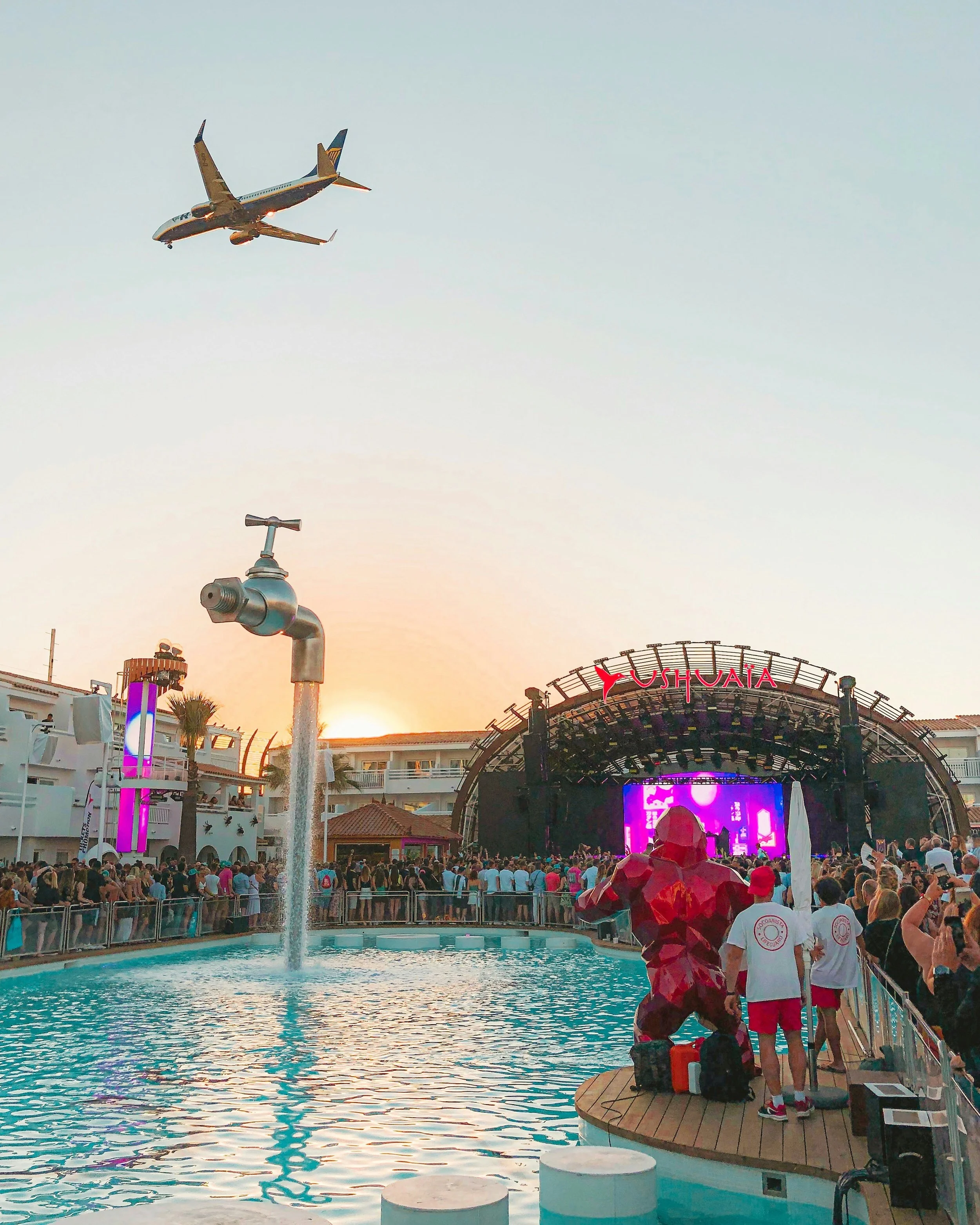 A large crowd gathered at a poolside concert venue during sunset with a stage labeled 'USHUAIA' in the background. An airplane is flying overhead in clear skies, and there are decorative structures and a fountain in the foreground.
