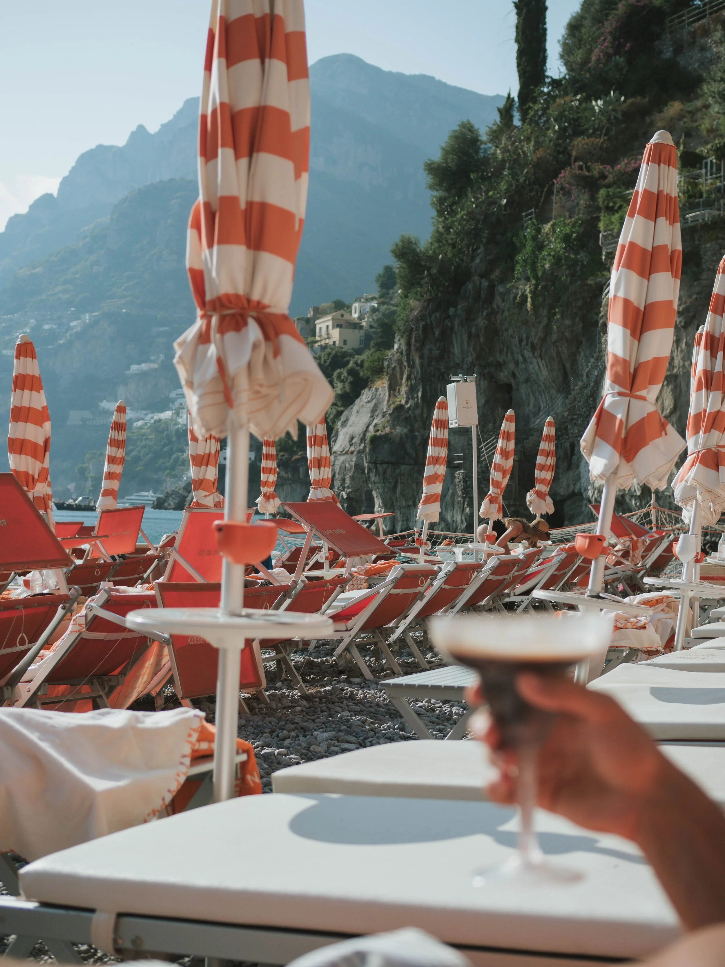 Beach scene with orange and white striped umbrellas and lounge chairs, mountains in the background, and a person holding a glass of drink in the foreground.