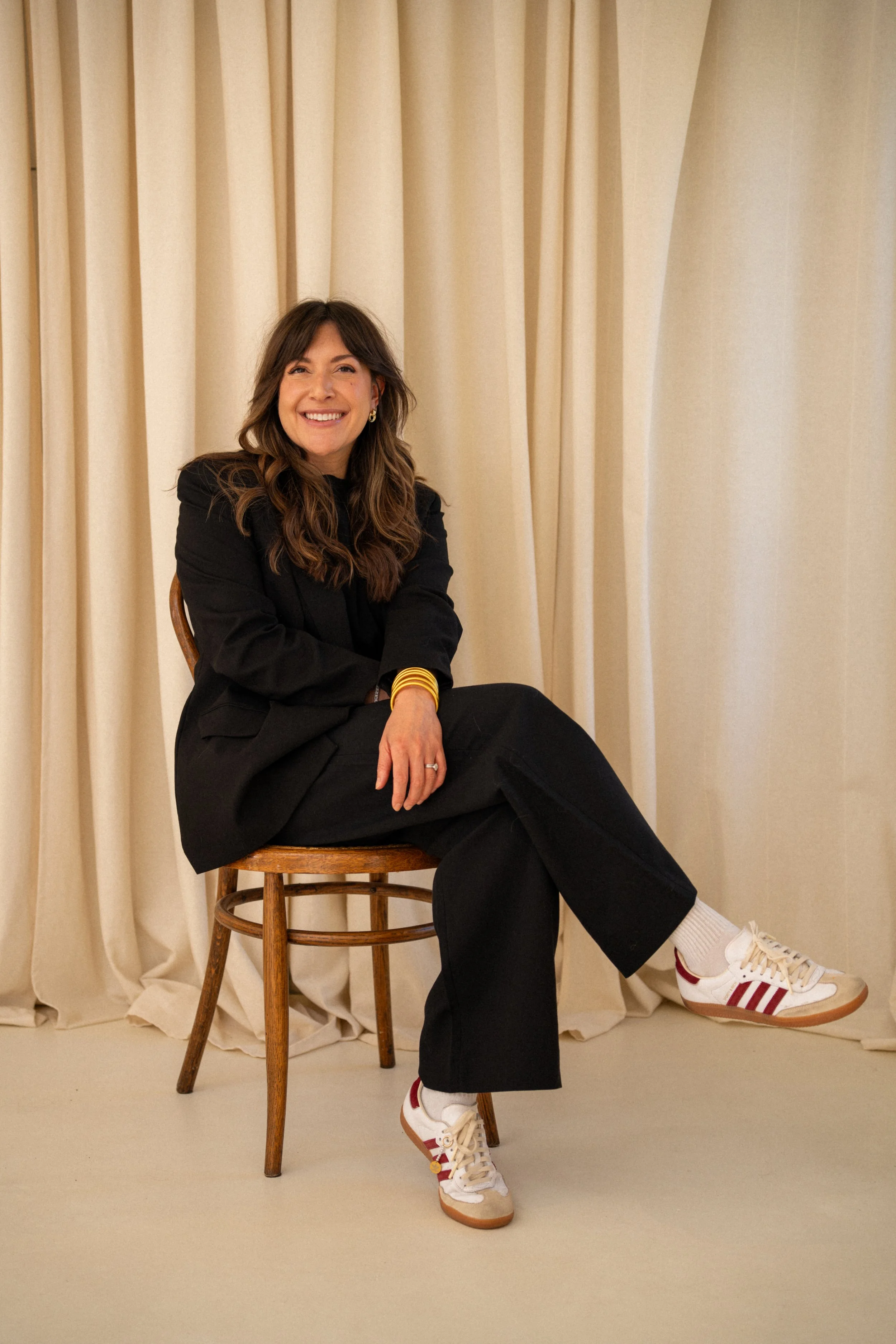 Smiling woman sitting on a wooden chair with her foot resting on another chair in front of a beige curtain background, wearing a black blazer, black pants, white sneakers with red stripes, and accessorized with gold jewelry.