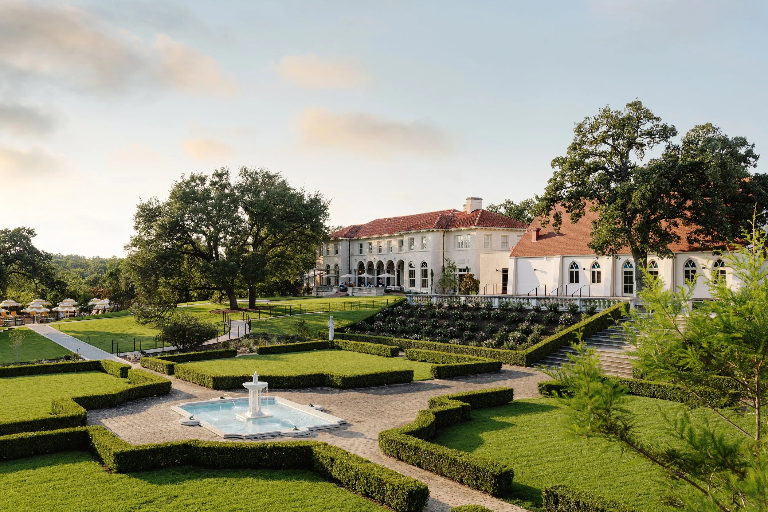 A large, luxurious estate with a white mansion featuring arched windows and a red-tiled roof, surrounded by well-manicured gardens, hedges, and a fountain, under a partly cloudy sky.