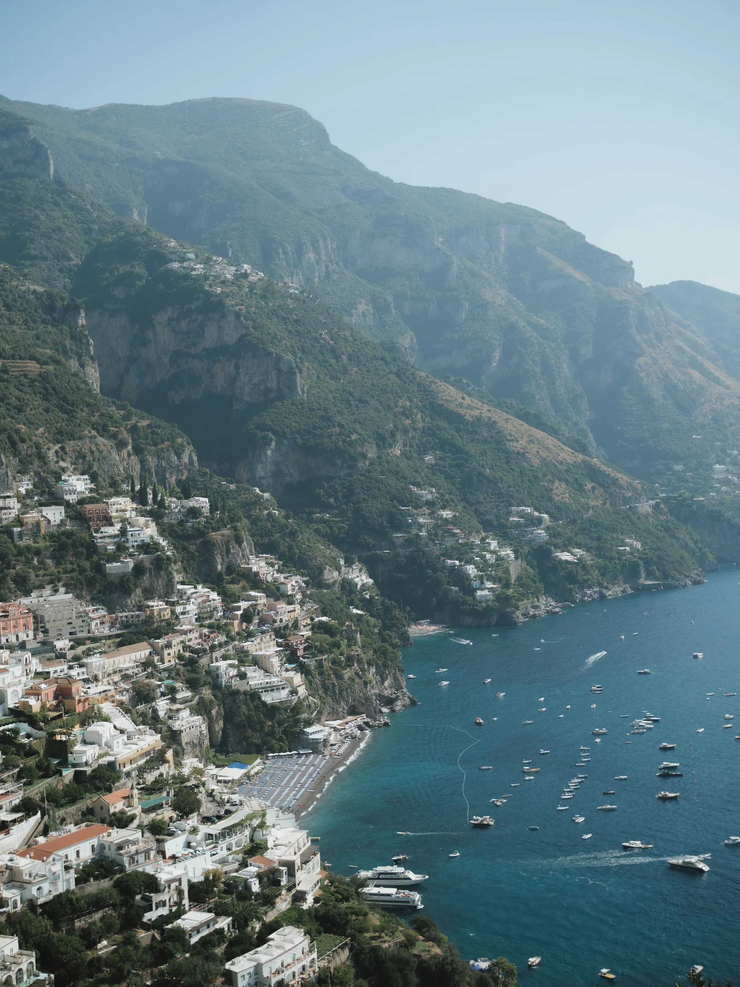 Coastal hillside town with white buildings, lush green mountains, and boats in the blue sea.