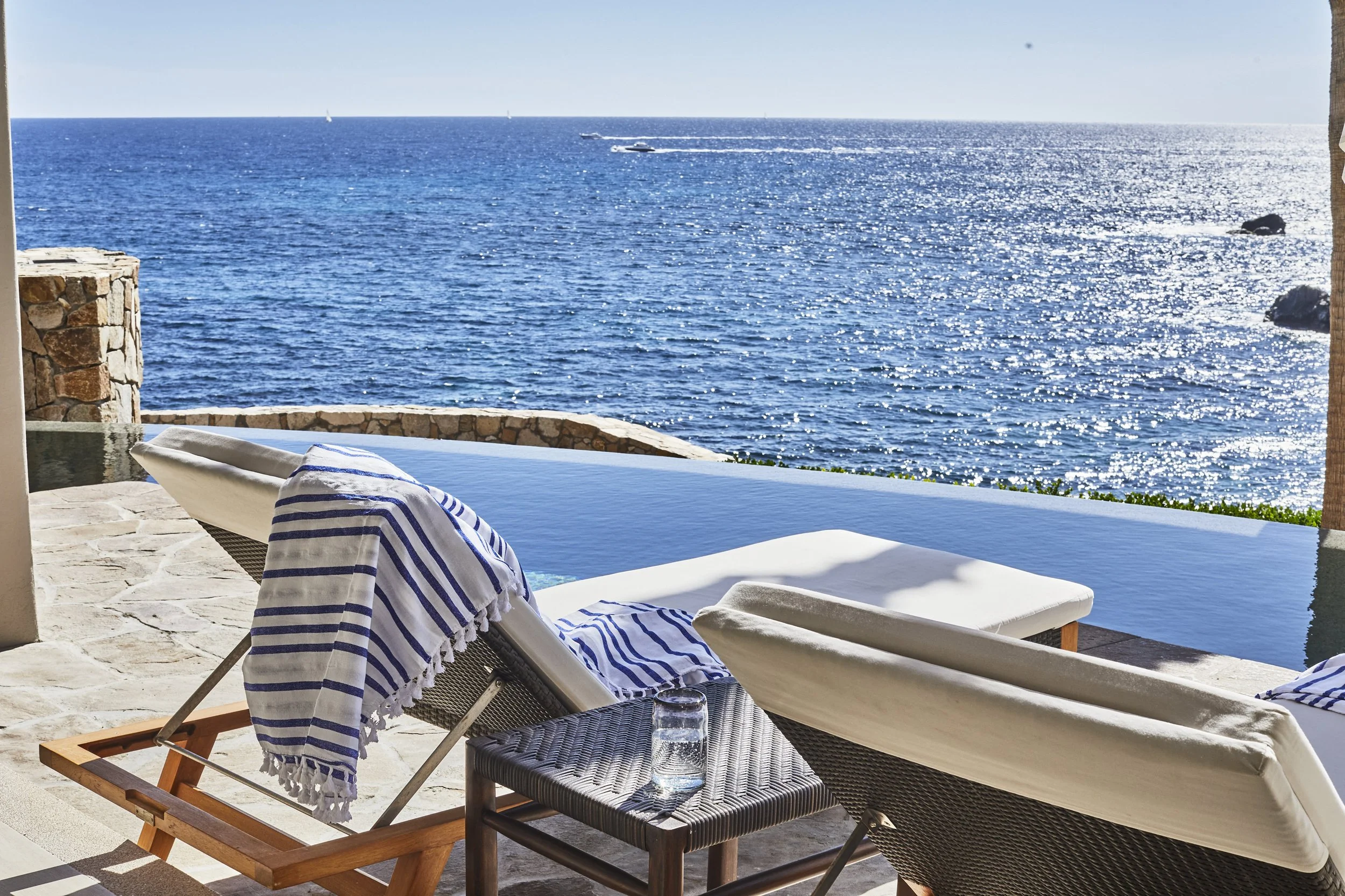 A view of a blue ocean with sailboats and two small boats leaving white wakes, seen from a shaded outdoor patio with lounge chairs and a glass of water.
