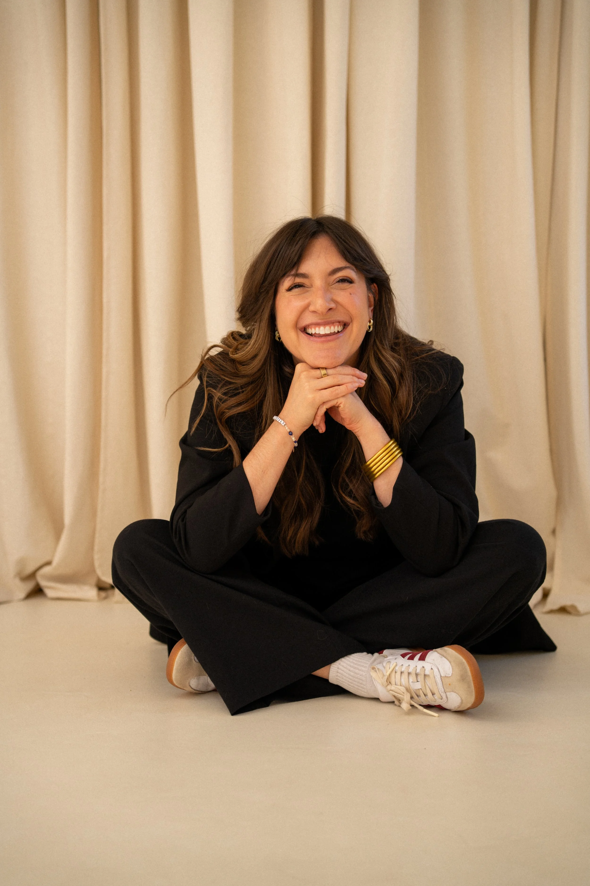 A woman with long brown hair smiling, sitting on the floor with her legs crossed in front of beige curtains, wearing a black suit, white sneakers with red stripes, and gold jewelry.