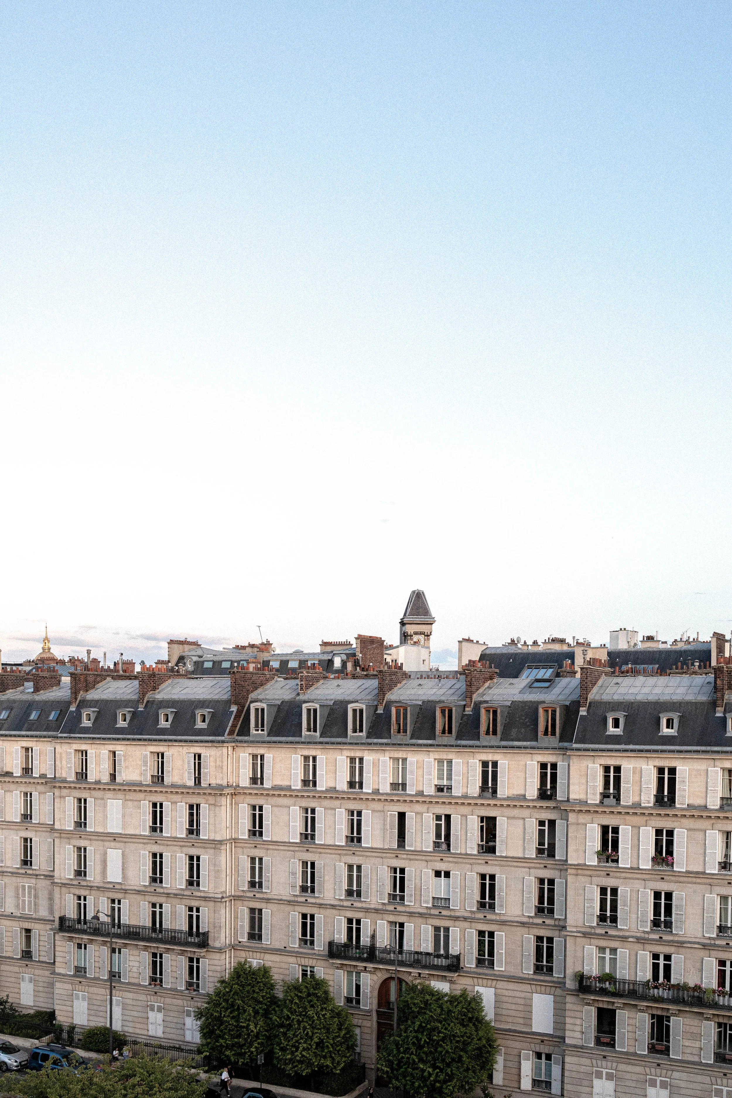 A view of a cityscape with a large building in the foreground, featuring multiple floors, windows, and balconies, and a green tree-lined street below. In the background, rooftops and a cloudy sky are visible.