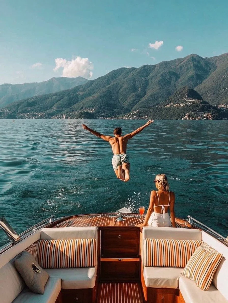 A man jumping off a boat into a large lake with mountains in the background, while a woman sitting on the boat's deck watches him holding a drink.