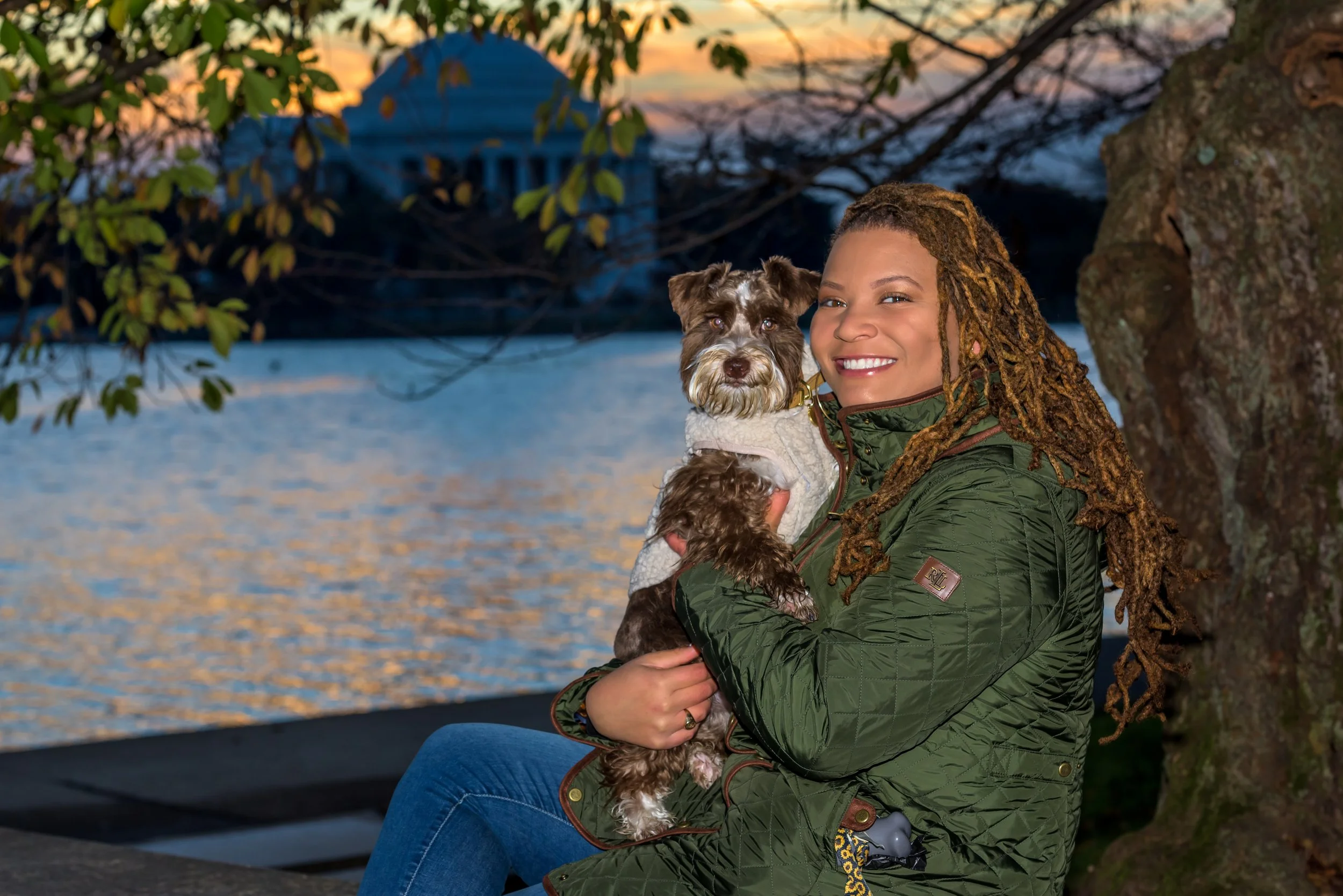 Portrait of a woman with long, braided hair smiling while sitting outdoors near a river, holding a small brown and white dog in her arms, with trees and a bridge in the background at sunset.