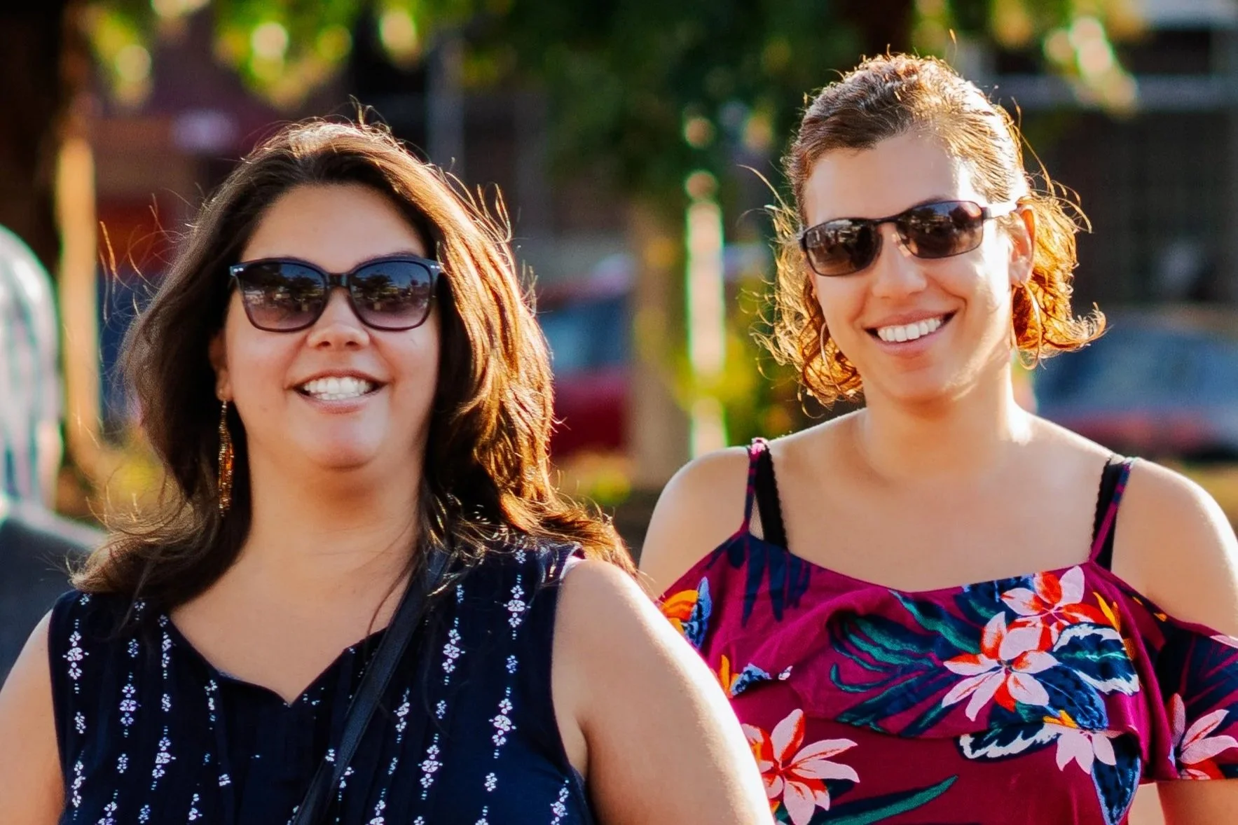 Candid portrait of two friends smiling outdoors, both wearing sunglasses and colorful summer dresses.