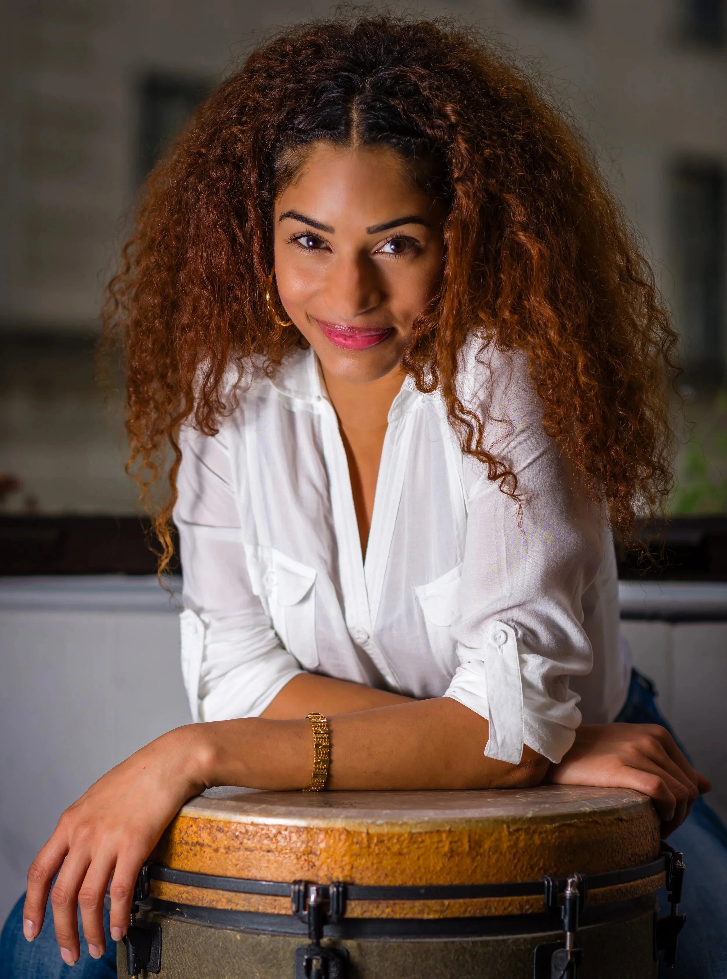 Portrait of a woman with curly hair wearing a white shirt, smiling and leaning on a drum, outdoors at night.