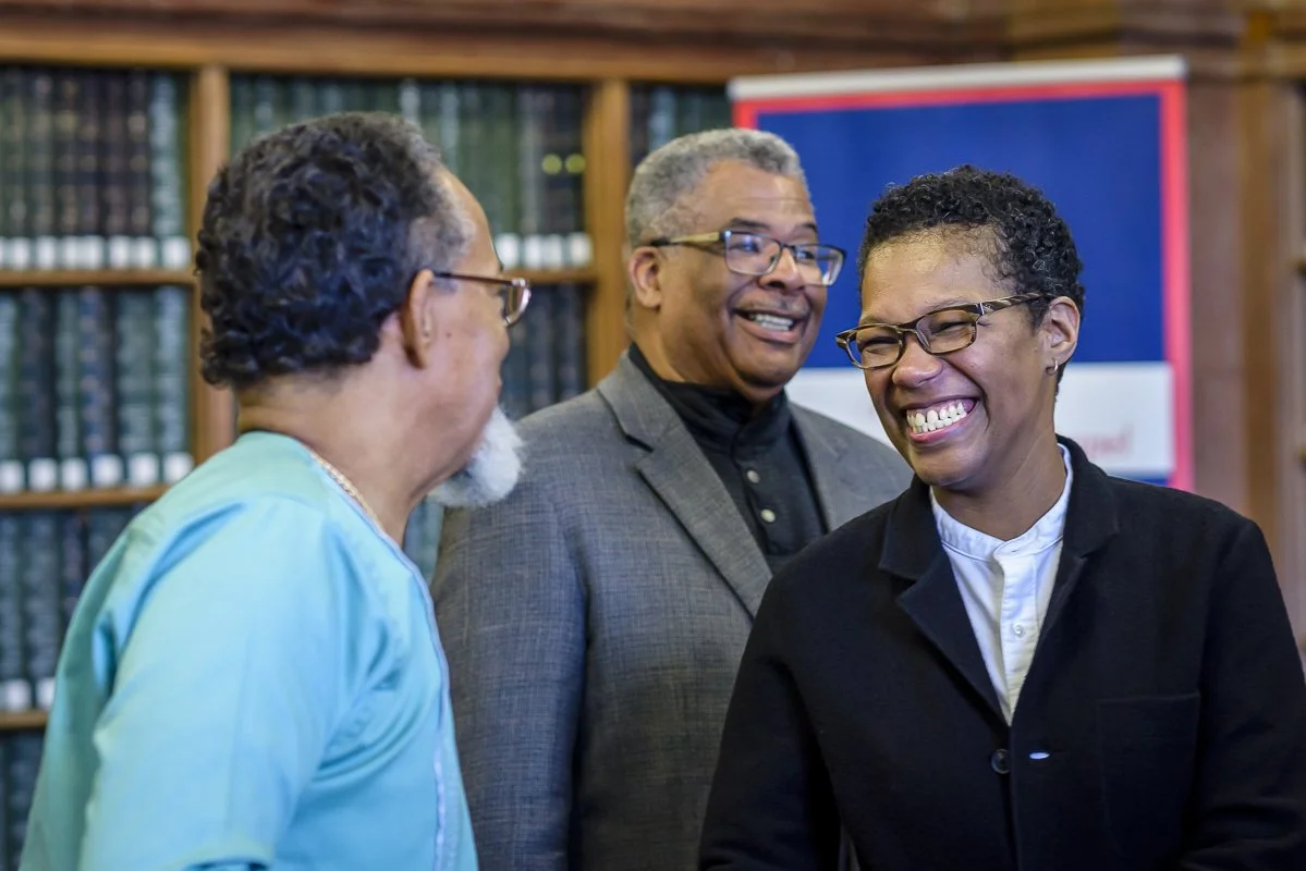 People smiling and chatting at an university event. The man on the left wears glasses and a blue jacket, the man in the middle wears glasses and a gray blazer, and the woman on the right wears glasses and a black jacket.