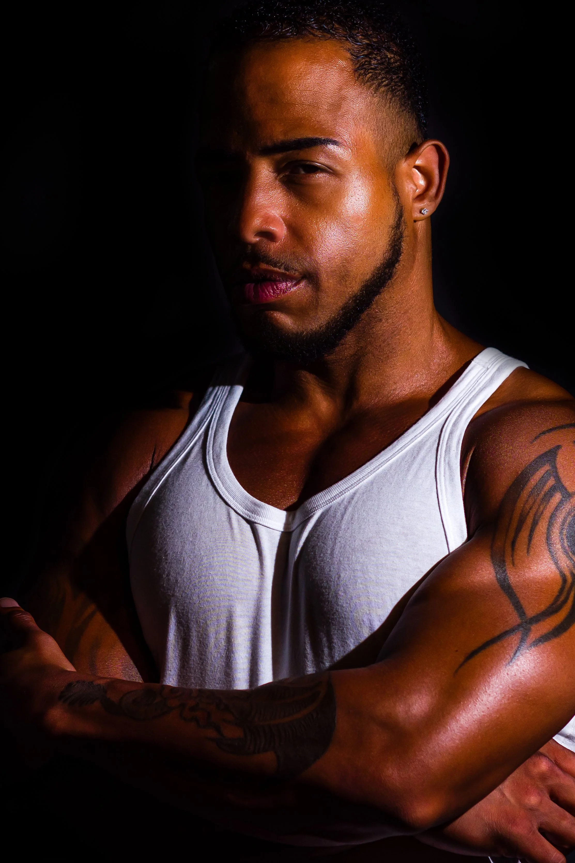 Portrait of a muscular man with tattoos on his arms, wearing a white tank top, posing with crossed arms against a dark background.