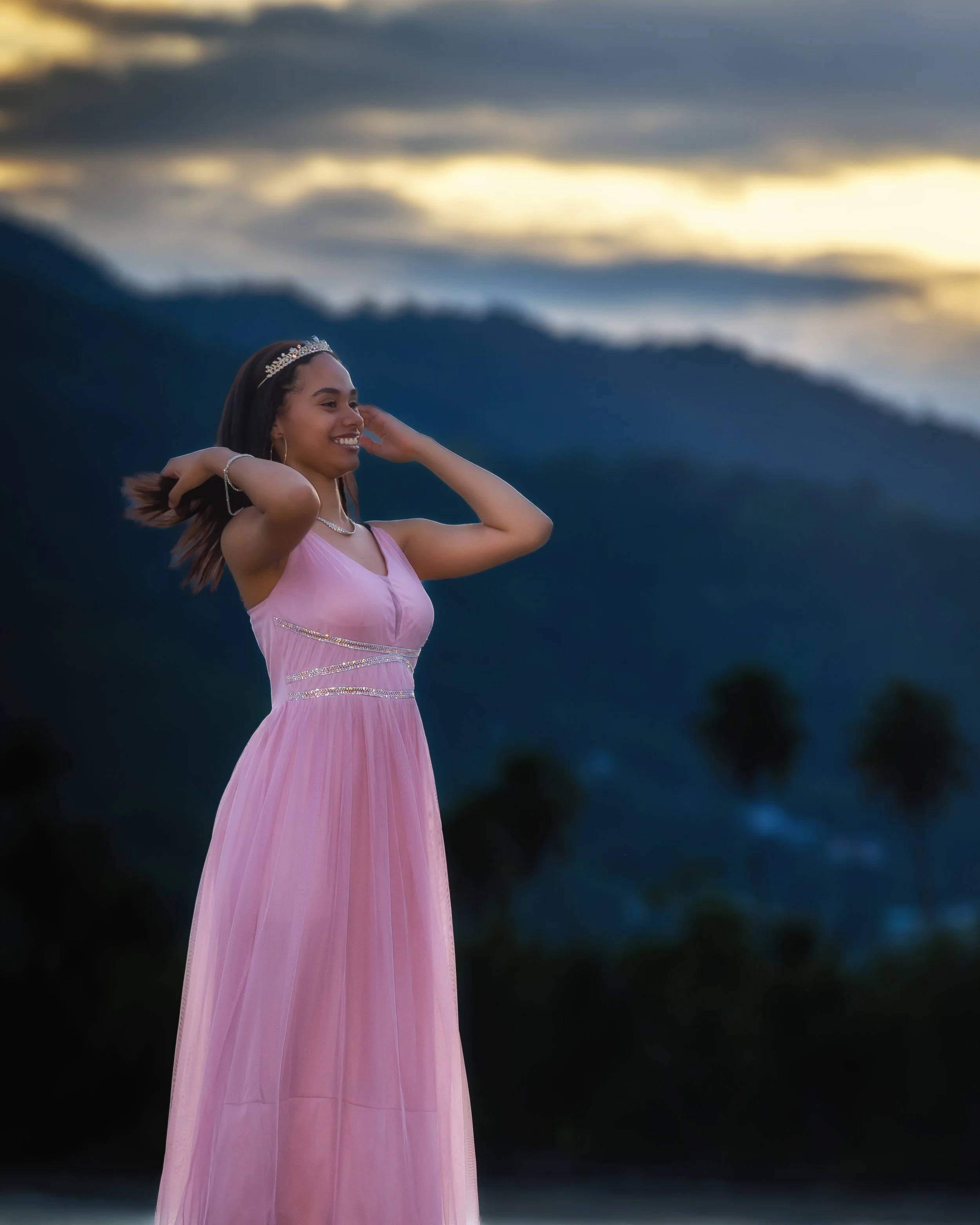 A young "quinceañera" woman in a pink gown with a tiara and jewelry, standing outdoors against a mountainous background during sunset or dusk, smiling and adjusting her hair as she celebrates her sweet 15.