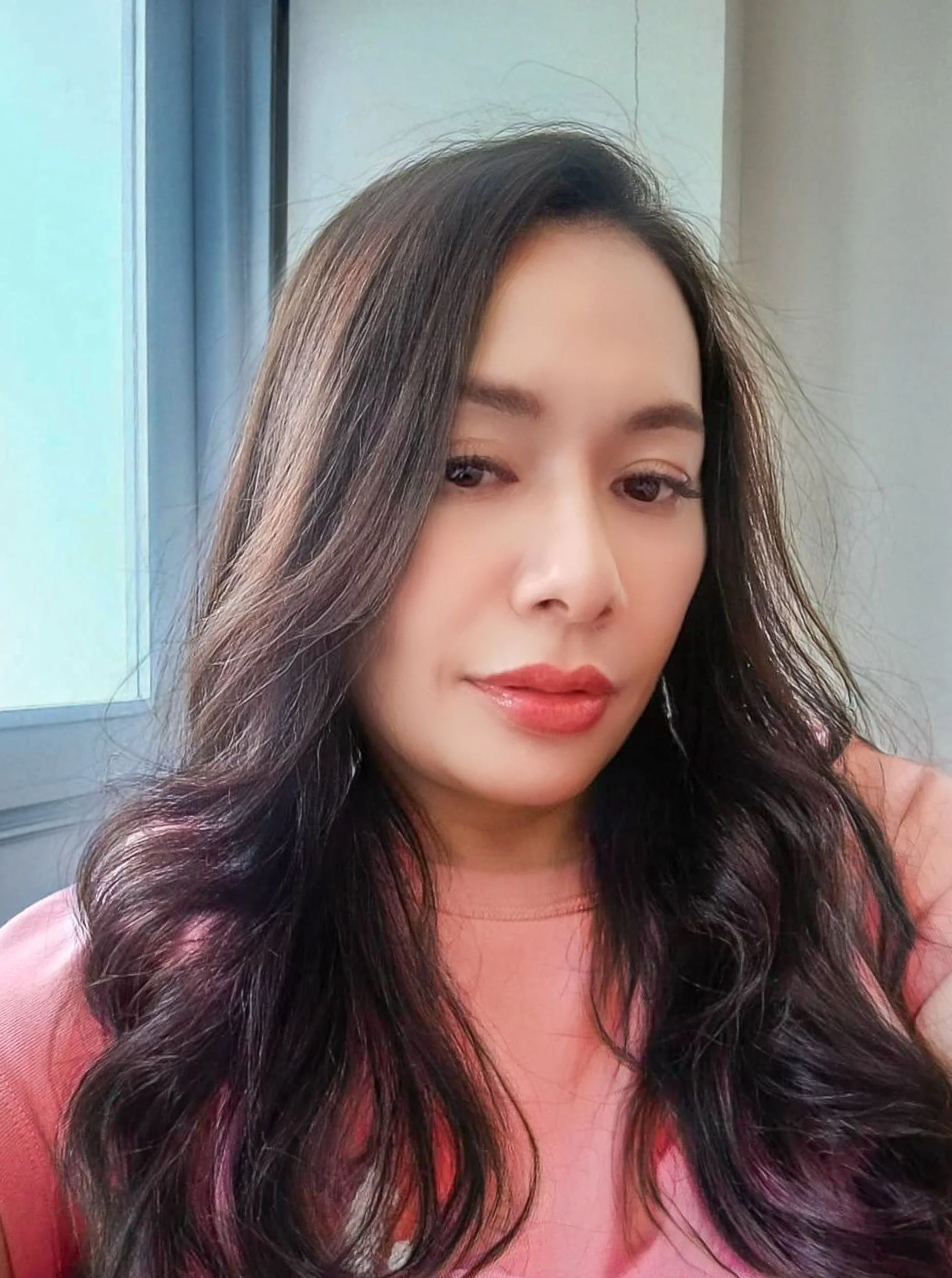 Portrait of a Thai woman with dark, wavy hair and wearing a pink top, sitting indoors near a window with blue curtains.