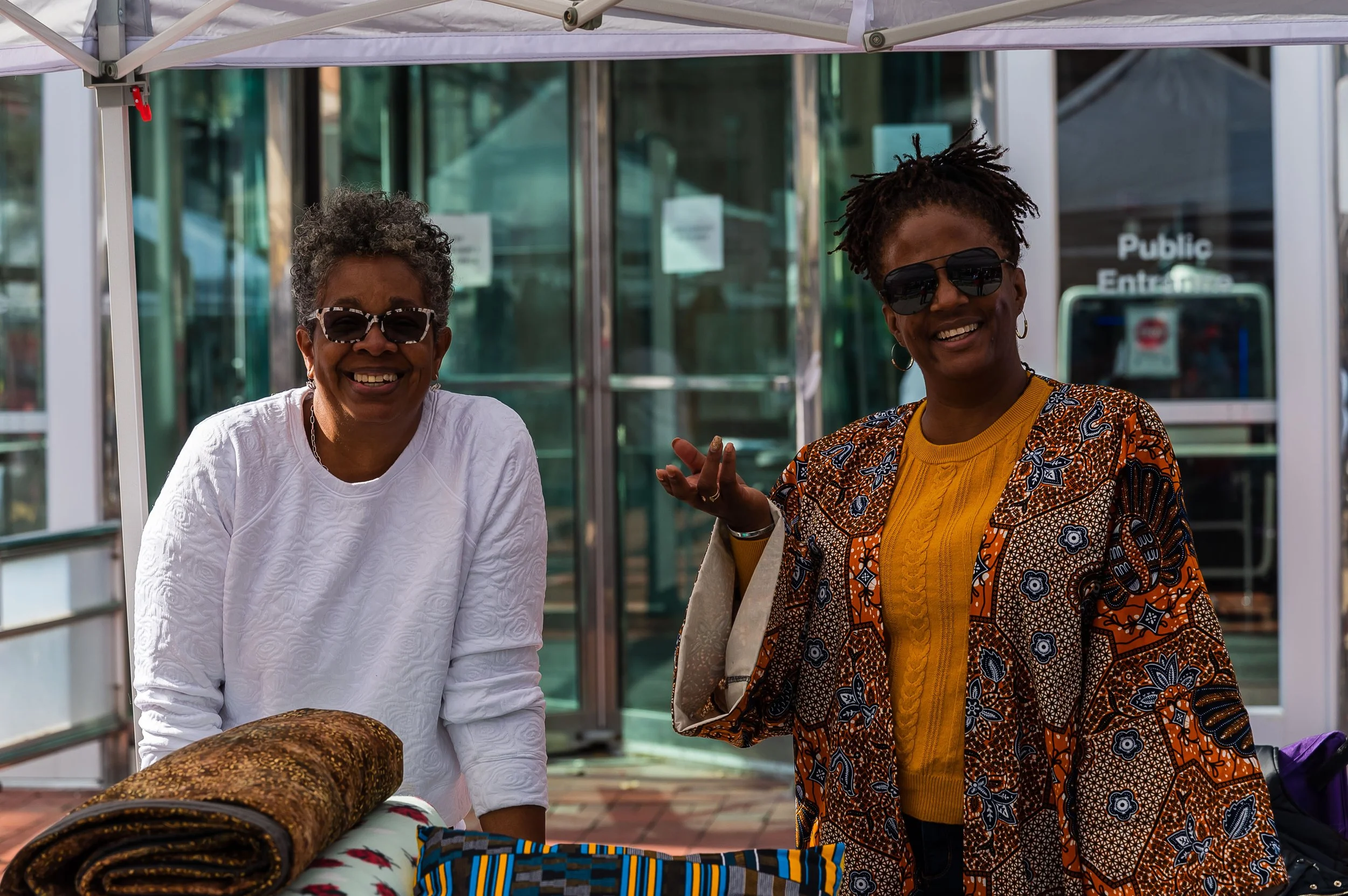 Two smiling vendors standing outdoors under a white canopy, with merchandise on a table in front of them, and a glass building in the background.