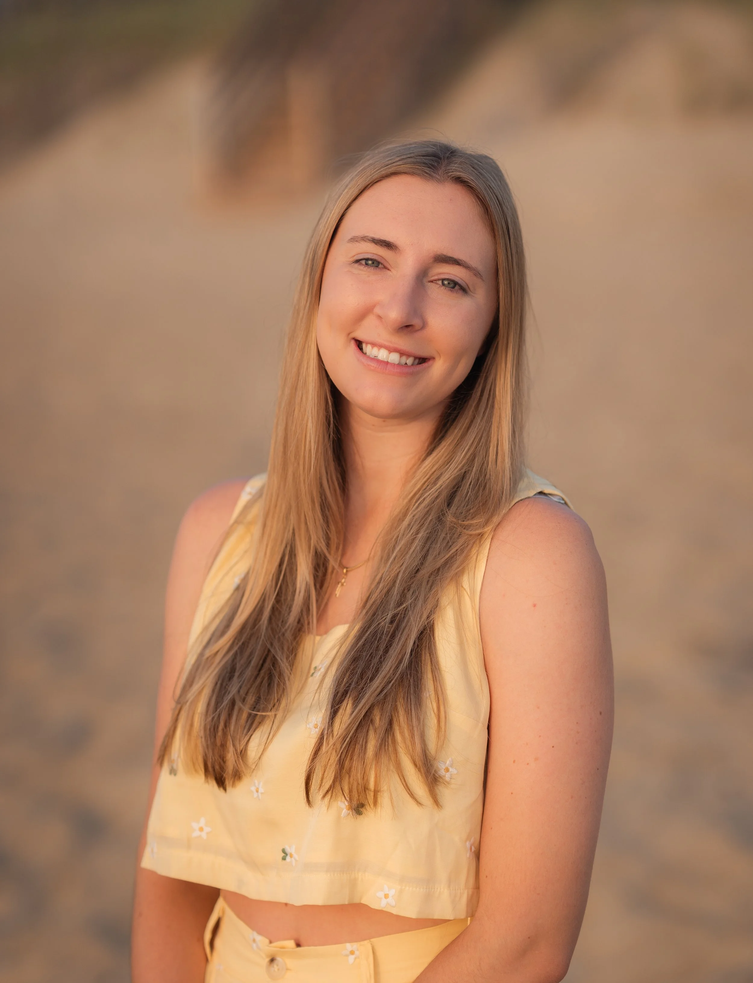 A young woman with long, blonde hair smiling outdoors at sunset, wearing a yellow sleeveless top with small white flowers, in a beach or sandy area.