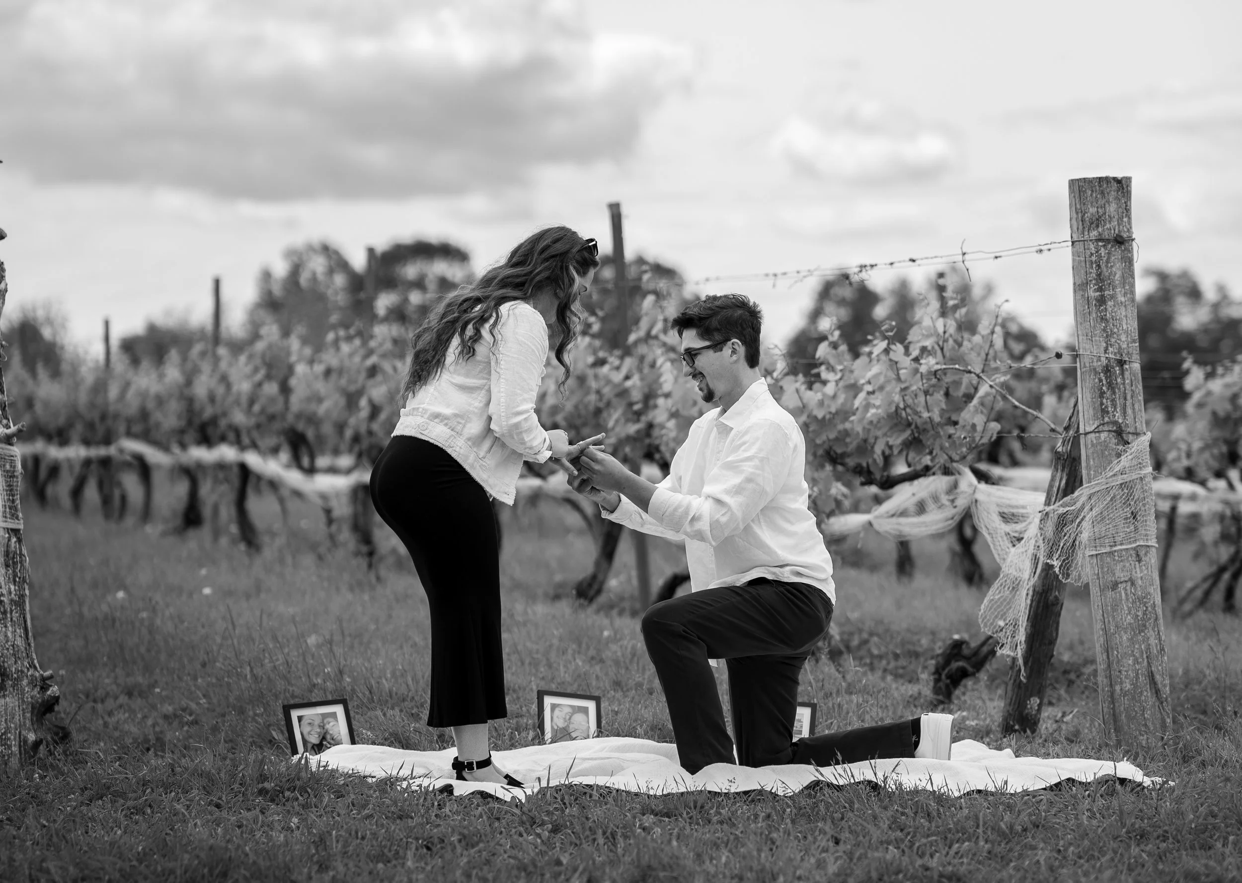 A man proposing marriage to a woman in a vineyard with framed photos laid on a blanket, during daytime in black and white.