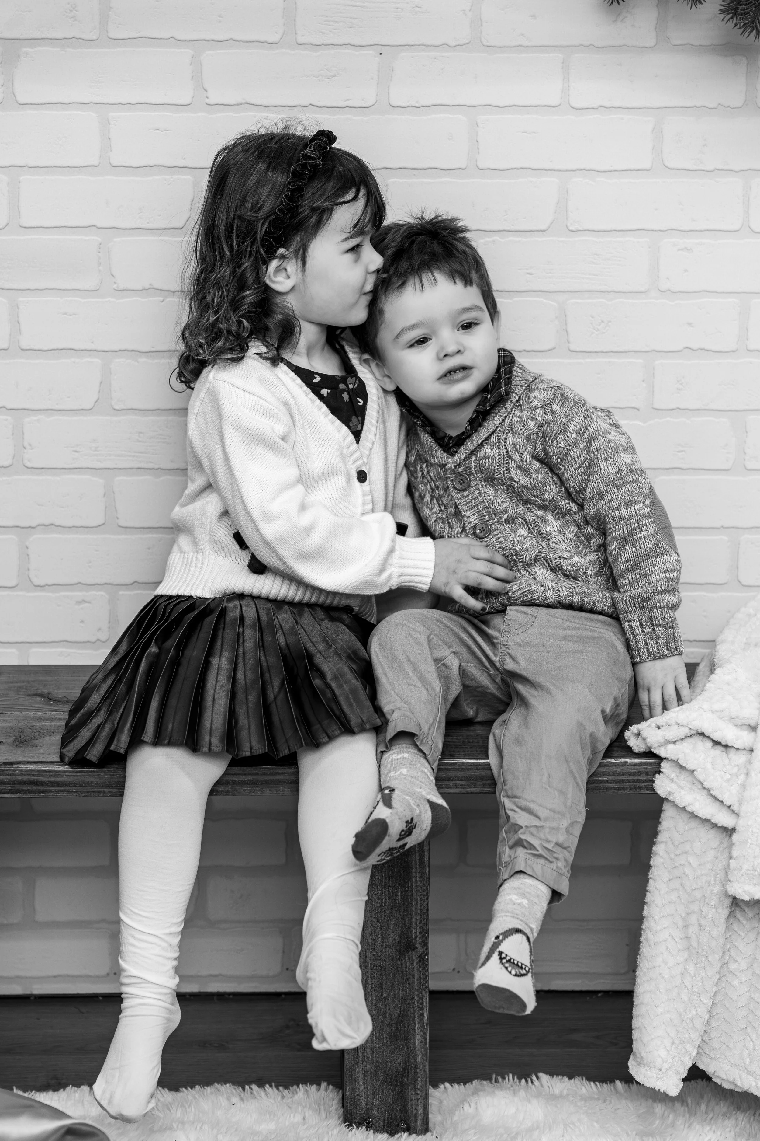 Two young children sitting on a wooden bench against a brick wall, the girl whispering to the boy, both wearing sweaters, the girl in a skirt and tights, and the boy in pants and socks.