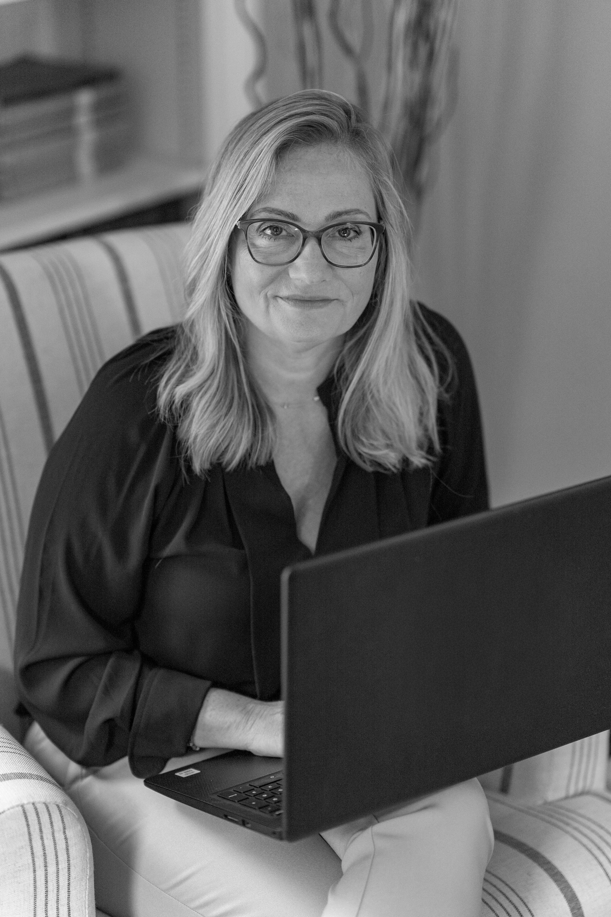 A woman with blonde hair, glasses, and a black blouse sitting on a striped armchair with a laptop on her lap, smiling at the camera.