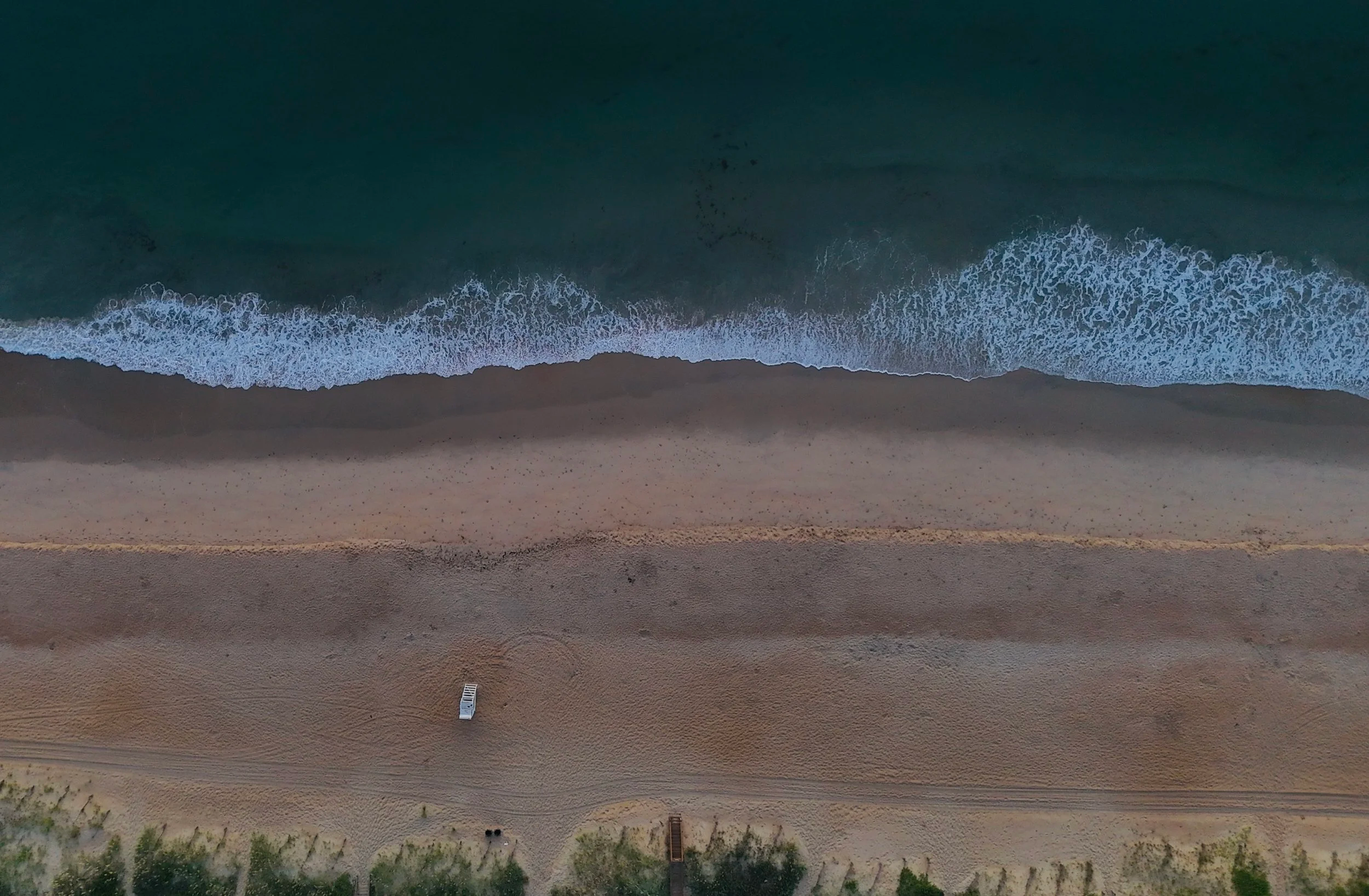 An aerial view of a sandy beach with waves crashing onto the shore and a single parked vehicle on the sand.