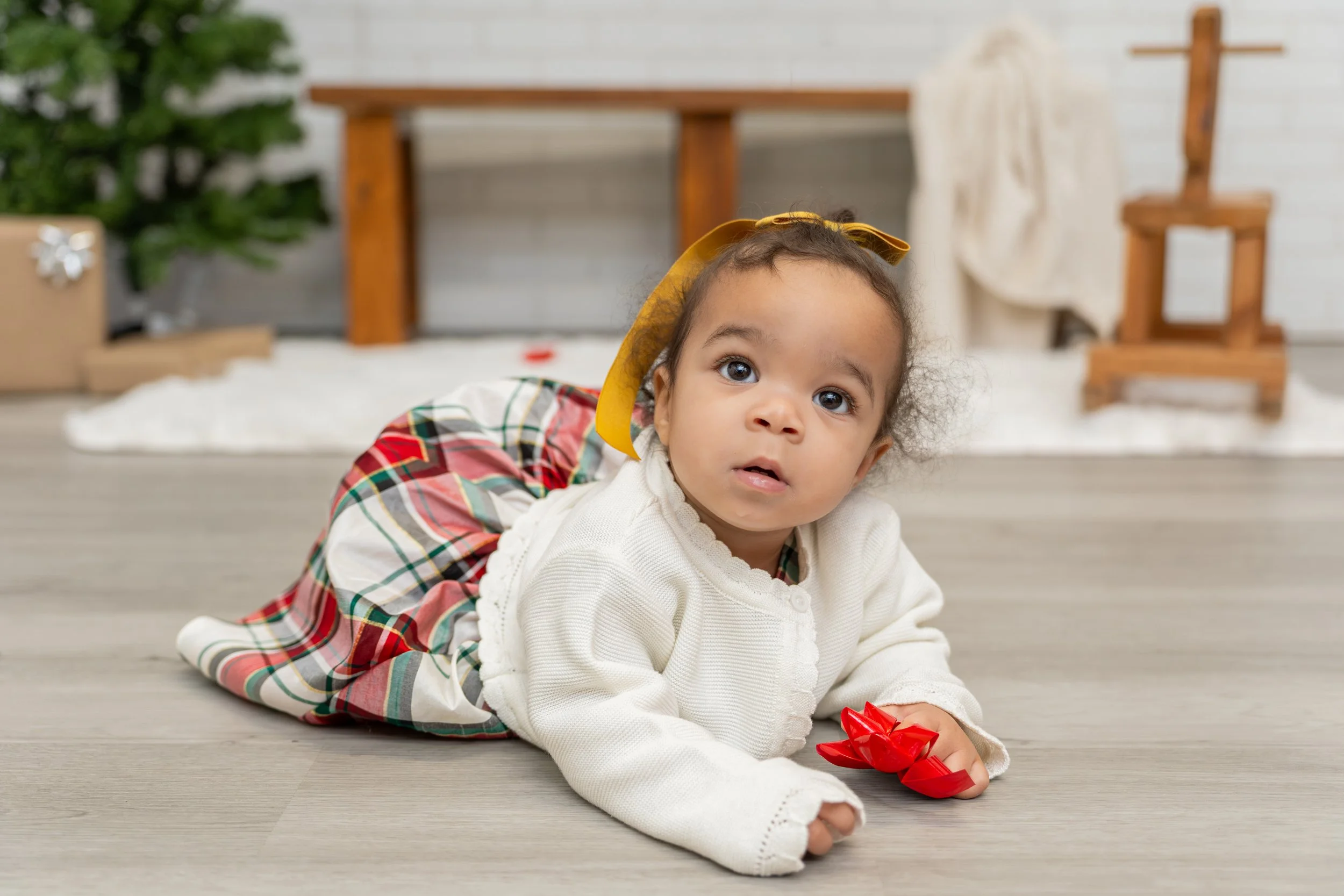 Young girl with a yellow ribbon in her hair, lying on a light-colored wooden floor, holding a red ribbon bow, in a room decorated for the holidays with a small Christmas tree, wrapped gifts, and a wooden chair in the background.
