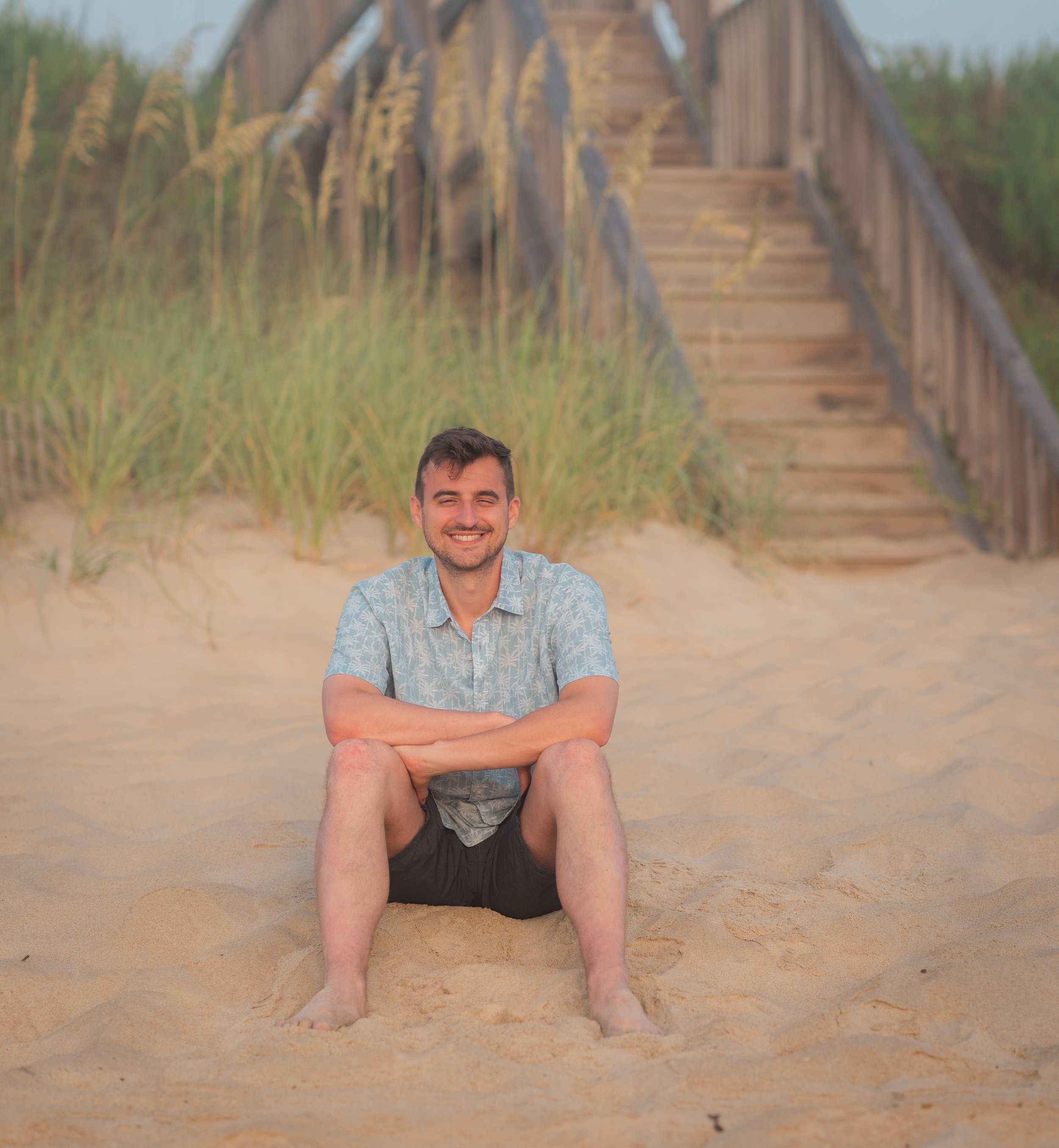 A man sitting on the sand at the beach, smiling with arms crossed, with a wooden staircase and beach grass in the background.