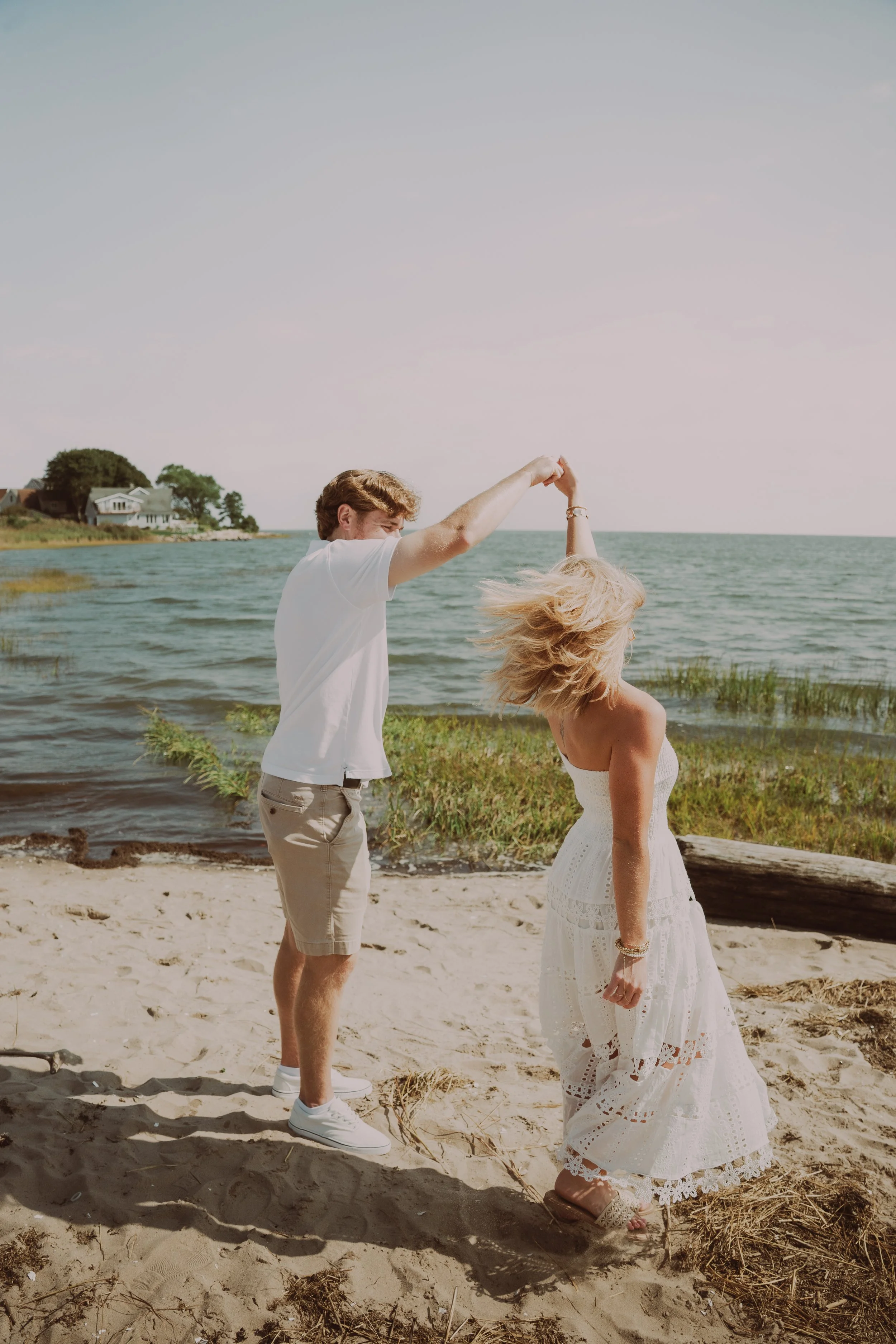 A couple dancing on a sandy beach near the water, with the man holding the woman's hand and spinning her around. The woman is wearing a white dress and the man is in casual clothing. There are houses and trees in the background along the shoreline.