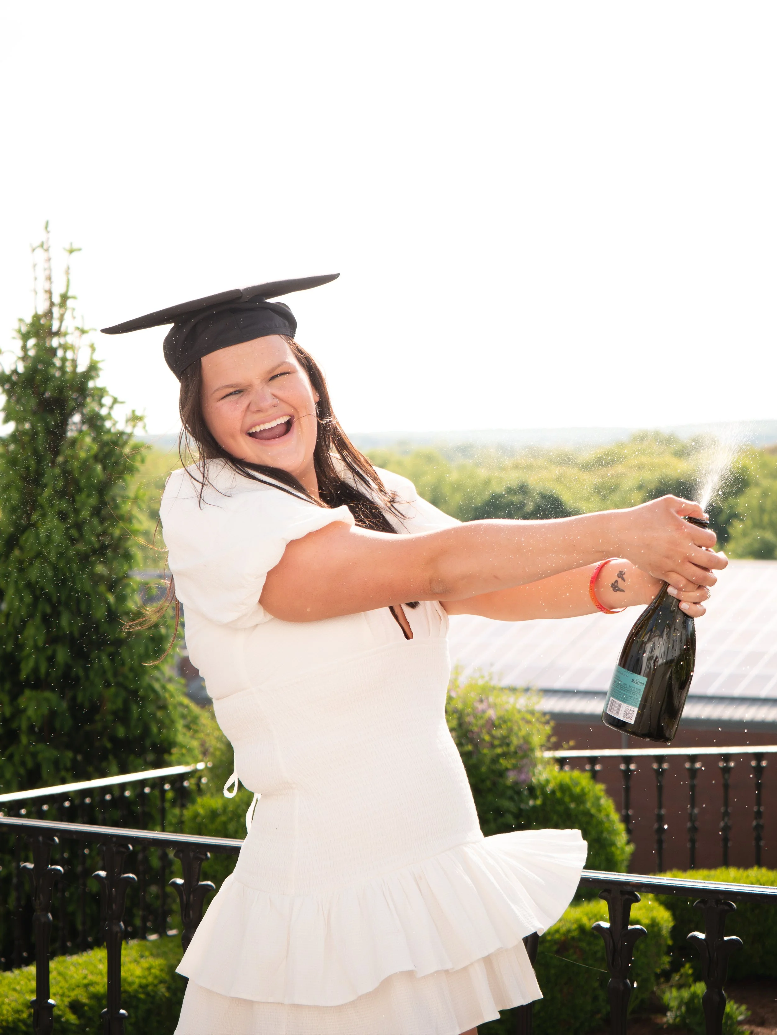 A woman wearing a white dress and black graduation cap celebrating outdoors with a bottle of champagne.