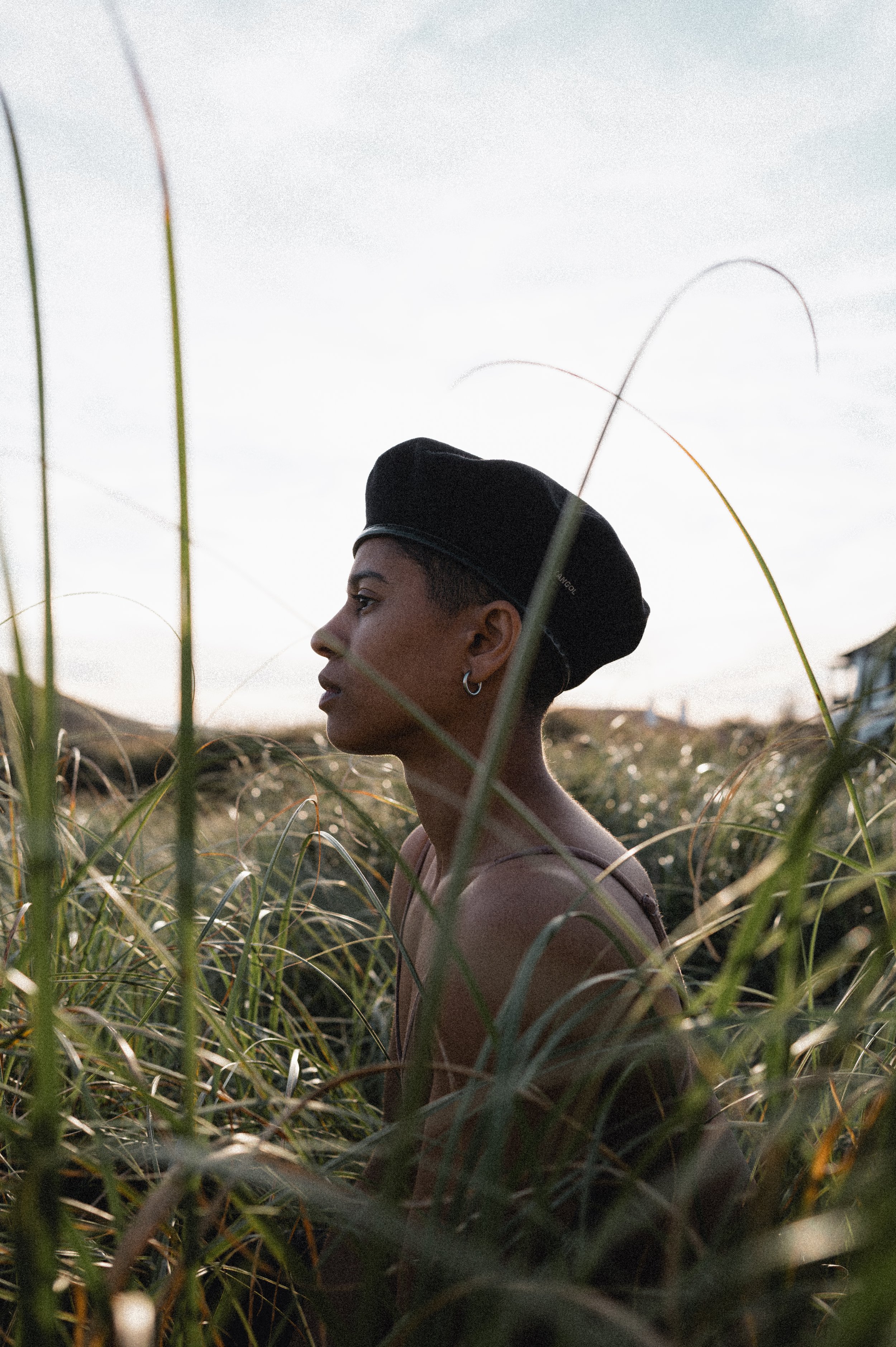 A young woman with dark skin and short hair, wearing a black hat, stands among tall grass during sunset or sunrise in an open field.