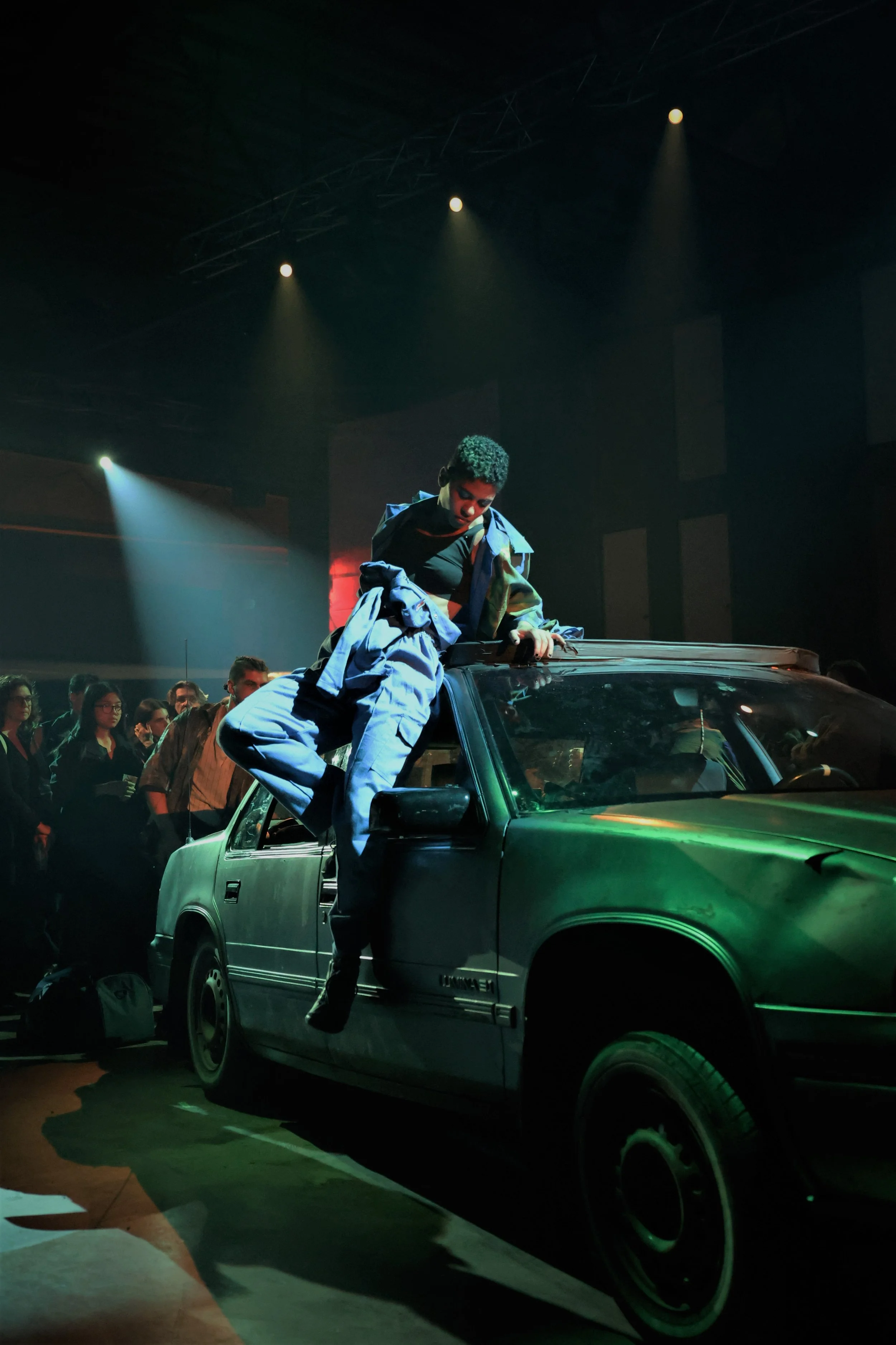 A young person with curly hair is sitting on the roof of a car, leaning forward and holding onto the edge. The car is green and parked indoors with a crowd of people around. Bright spotlights are shining from above, creating a dramatic atmosphere.