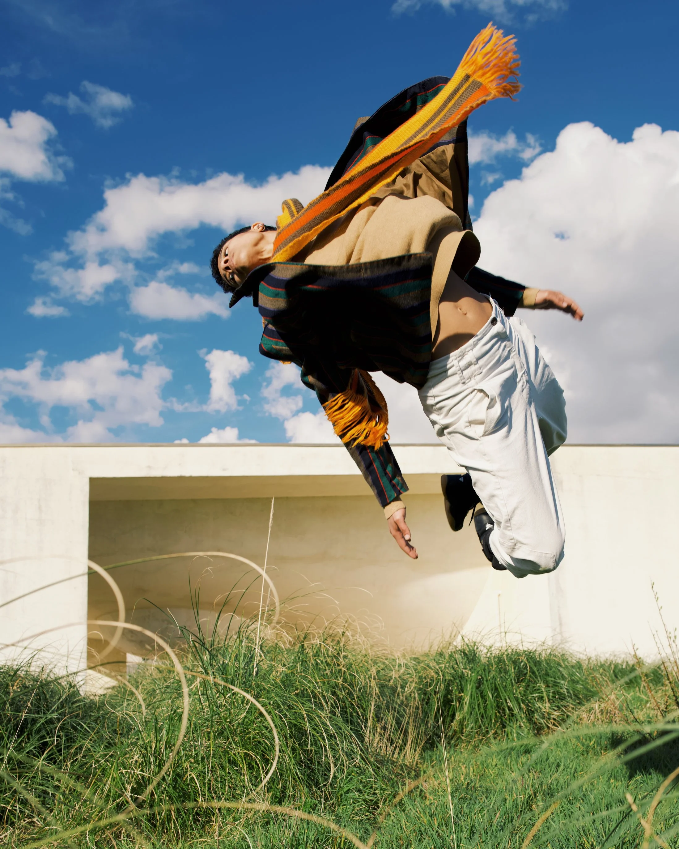 A person in casual clothing, including a beige jacket, white pants, and black shoes, is leaping or falling with arms outstretched against a blue sky with scattered clouds. They are outdoors near a white structure, with green grass and plants at the bottom of the image.