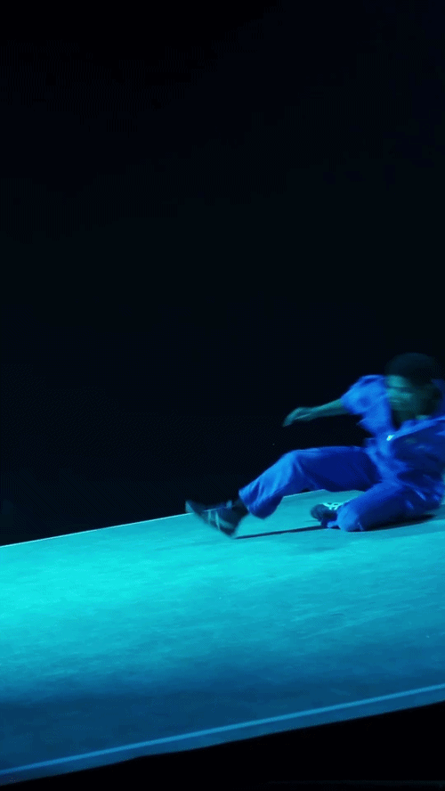 A person in a blue outfit appears to fall or slide on a stage with a dark background, in a dynamic pose.