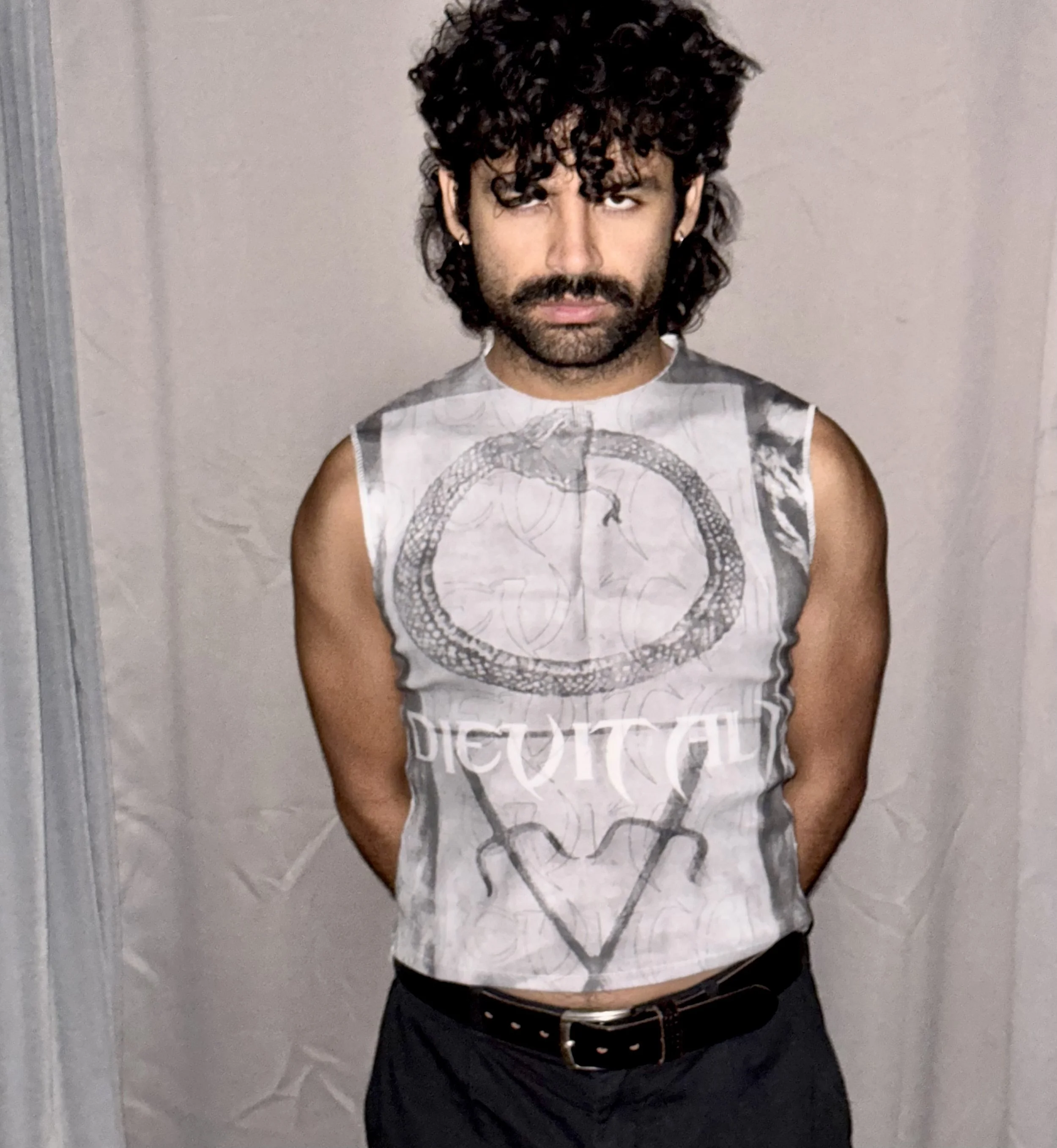 A man with curly dark hair, a mustache, and beard, wearing a sleeveless shirt with a snake and text design, standing against a plain wall.