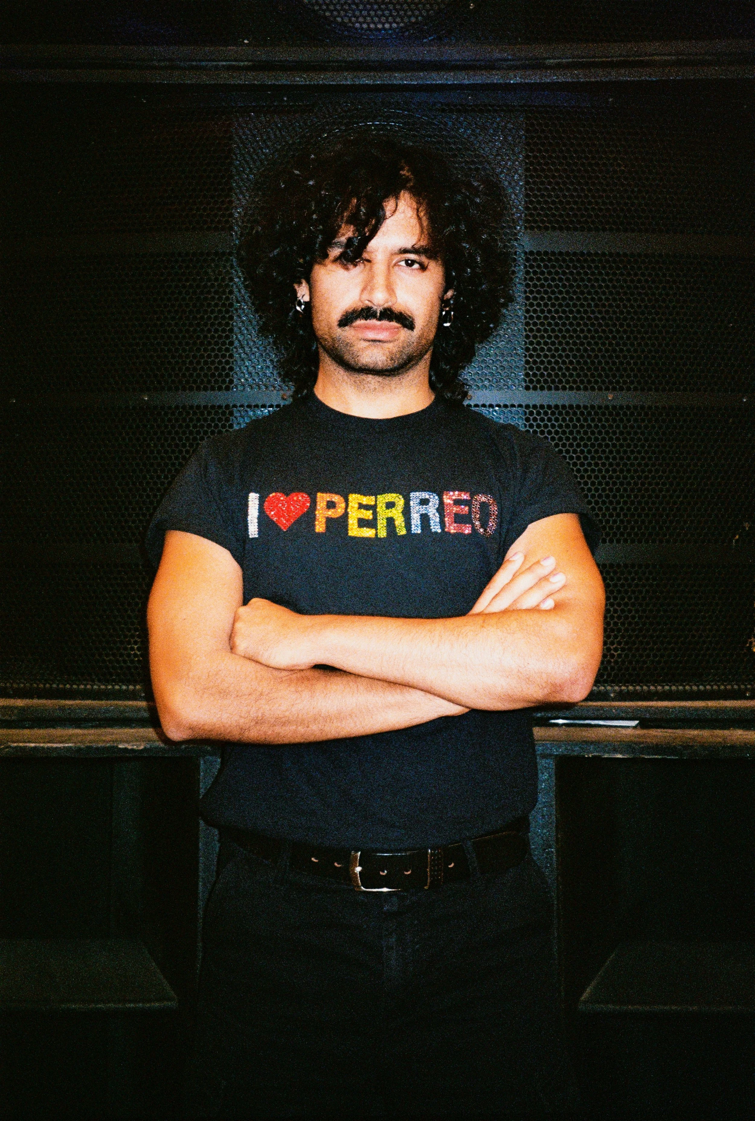 A man with curly black hair and a mustache stands with his arms crossed, wearing a black t-shirt that says "I love PERREO" with a red heart and multicolored letters.