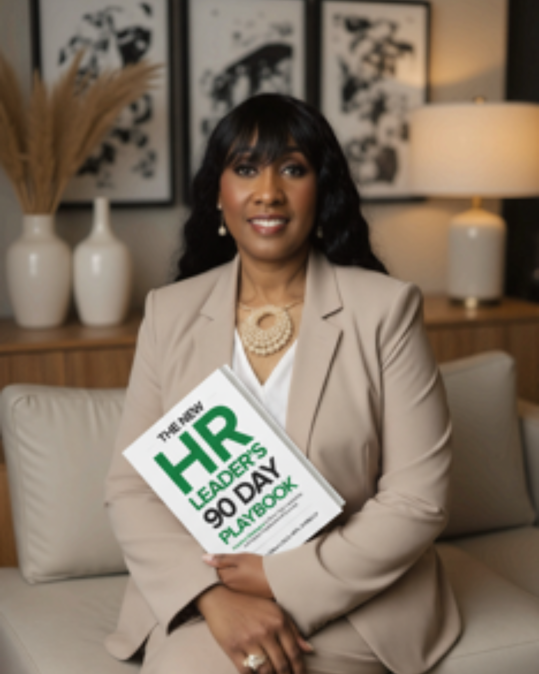 A woman in beige business attire sitting on a couch, holding a booklet titled 'The Top HR Leaders of 90 Day Playbook'.