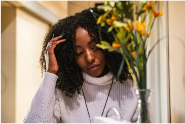 Woman with dark curly hair and light turtle neck, eyes lowered, hand resting on her head besides flowers contemplating change