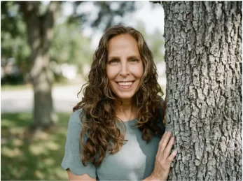 Smiling woman with long curly hair standing beside a large tree outdoors