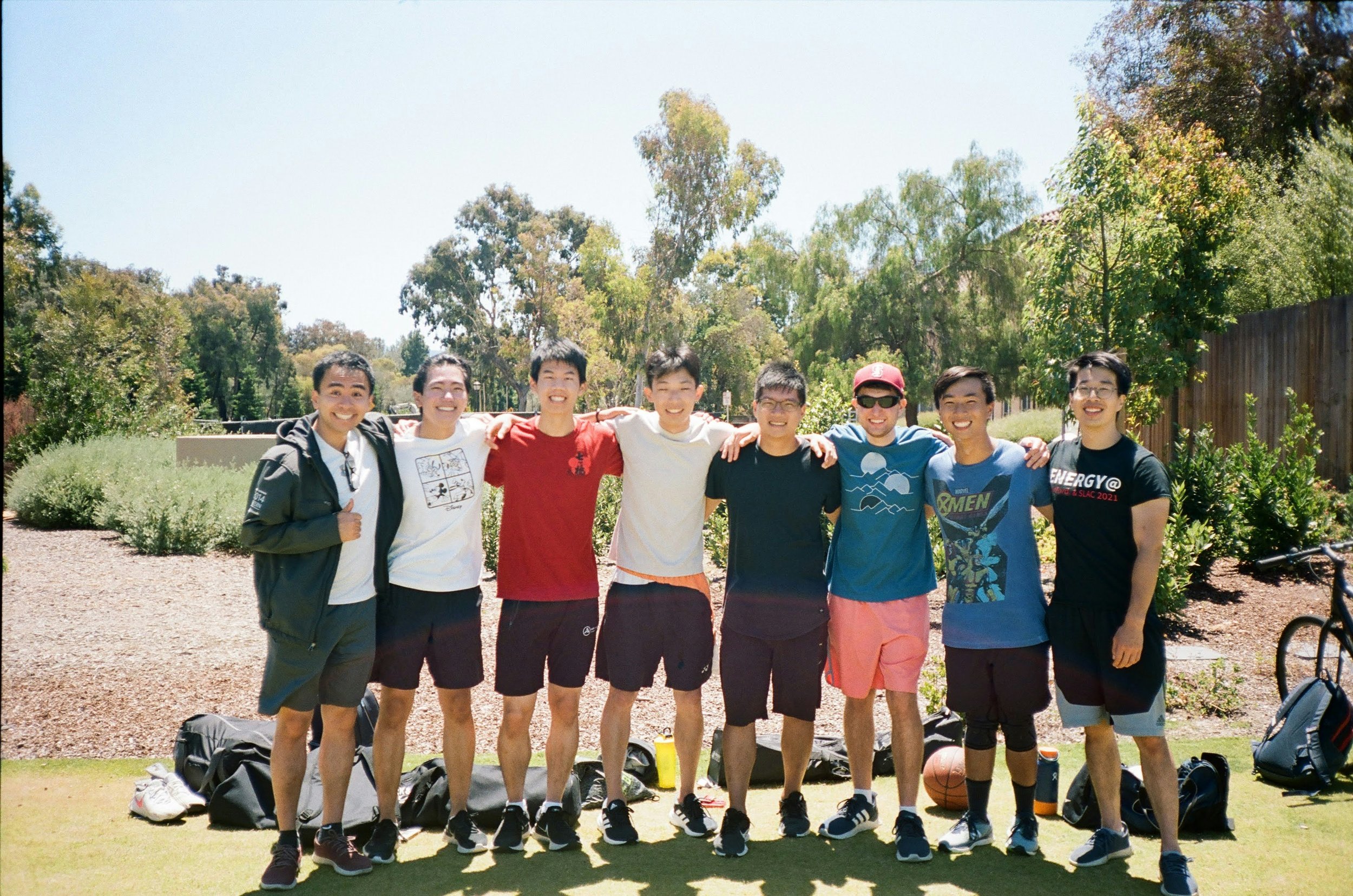 A group of nine young men standing outdoors with their arms around each other's shoulders, smiling at the camera. They are dressed casually, with some in t-shirts, shorts, and sneakers. The background features trees, bushes, and a wooden fence, with a sunny sky.
