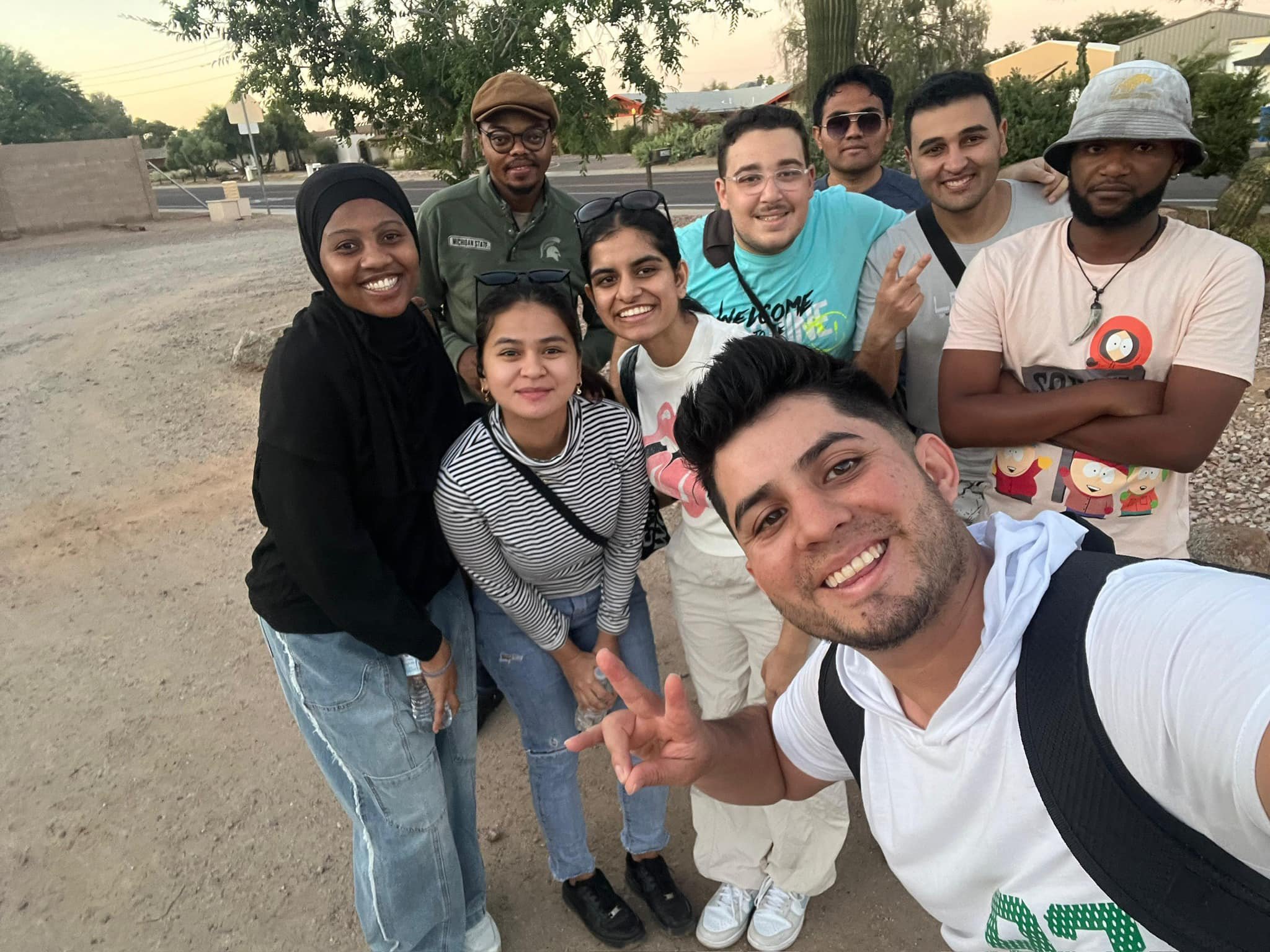 A group of nine diverse friends smiling and posing for a selfie outdoors during sunset.