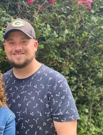 A smiling man with a beard wearing a baseball cap and a navy T-shirt, standing outdoors with green foliage and pink flowers in the background.