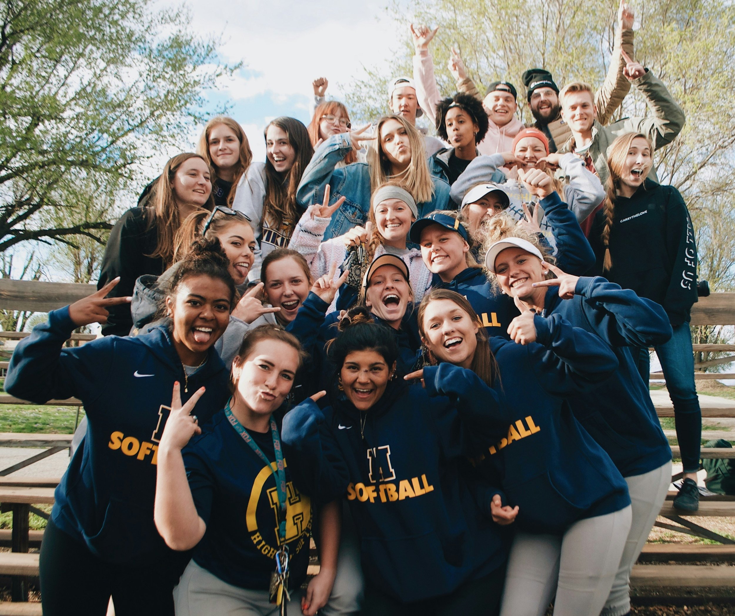 Group of young women and men gathered outdoors on bleachers, smiling and making playful gestures, some wearing navy blue softball jerseys with yellow letters.