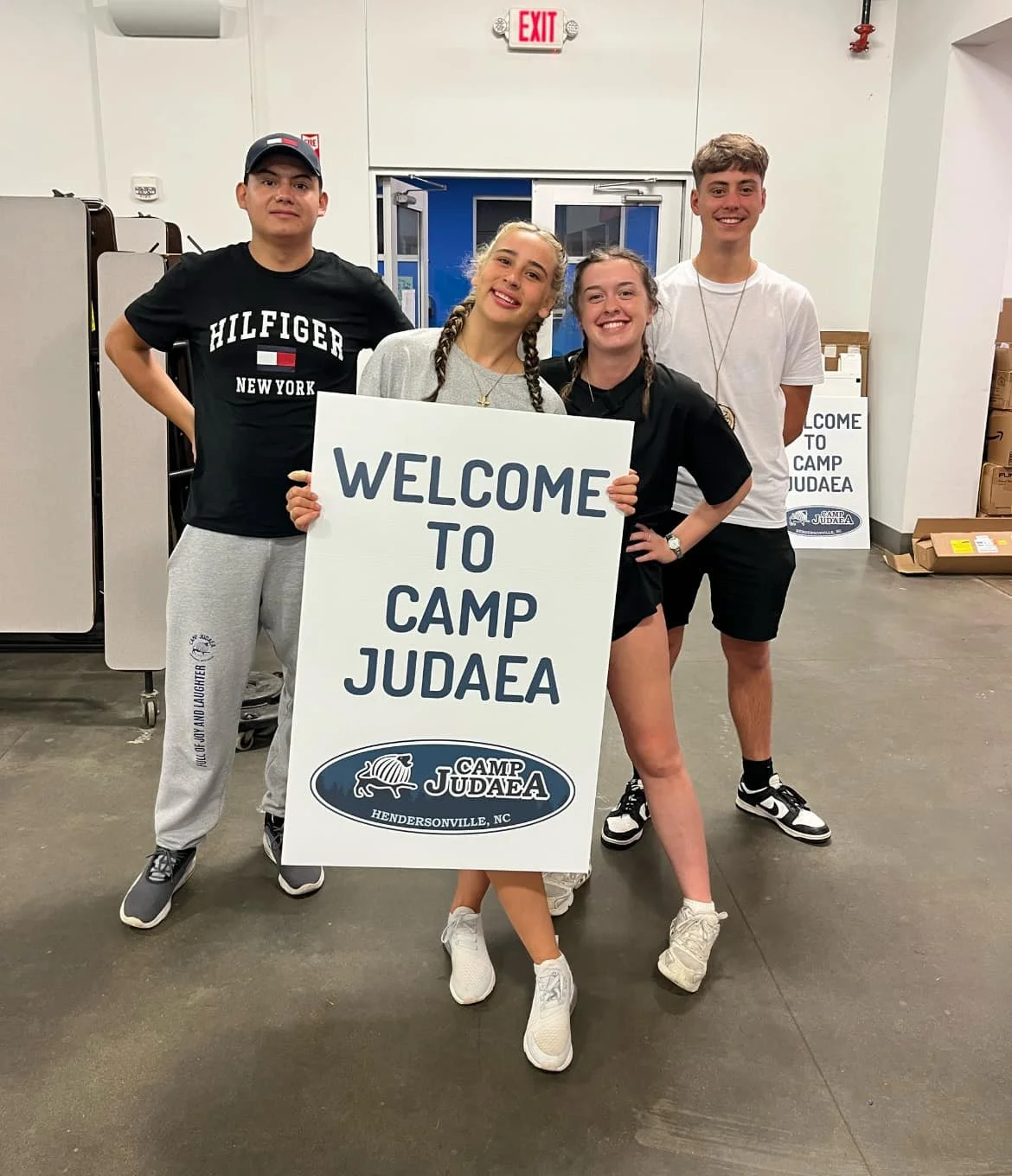 Four teenagers standing inside a building, holding a large sign that reads 'Welcome to Camp Judaea'. They are smiling and wearing casual clothes, with some wearing shorts and sneakers. The background includes a sign that says 'Welcome to Camp Judaea' and an exit door.