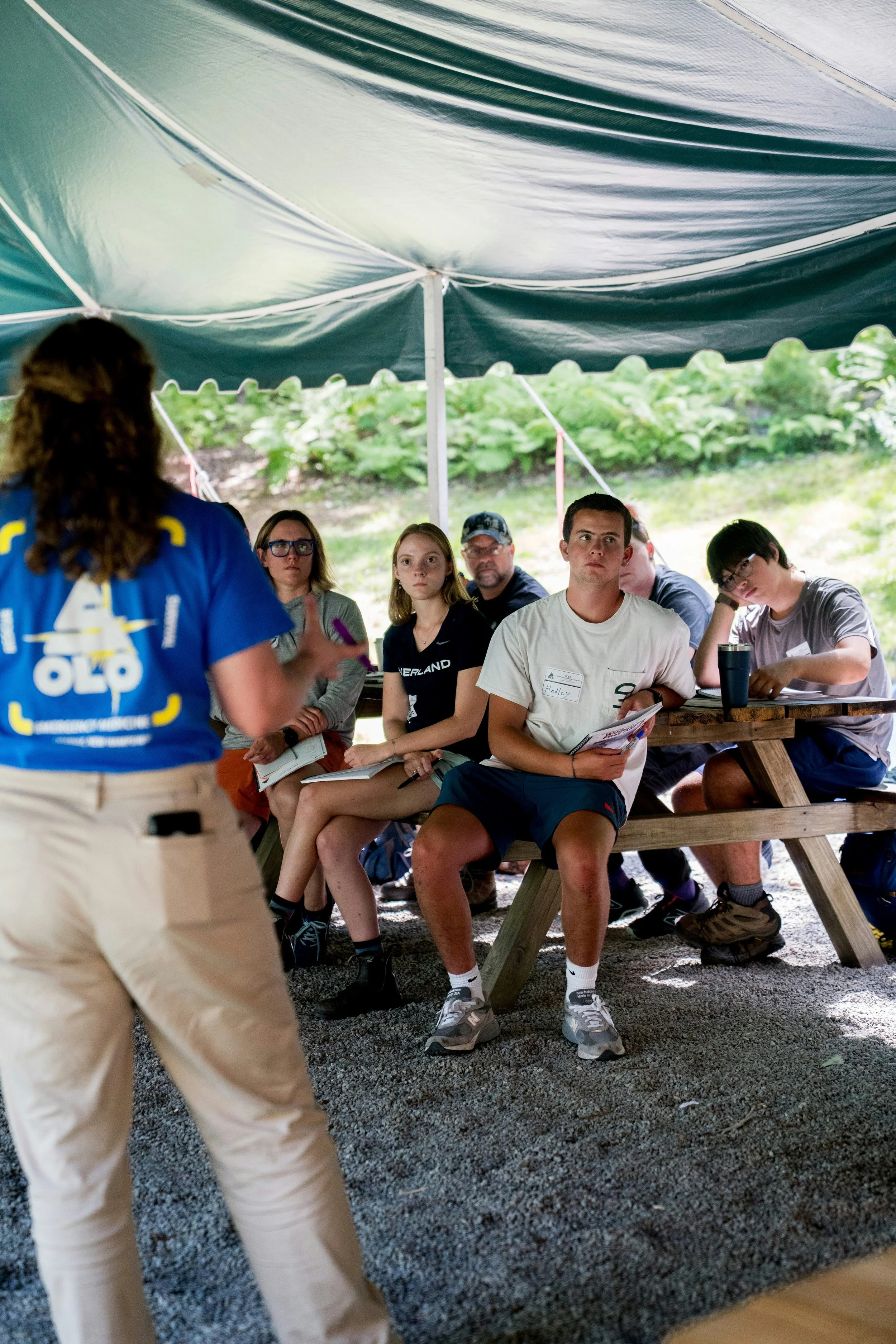 A group of young people sitting at a picnic table inside a large outdoor tent, listening to a woman standing in front of them, giving a presentation or speech.
