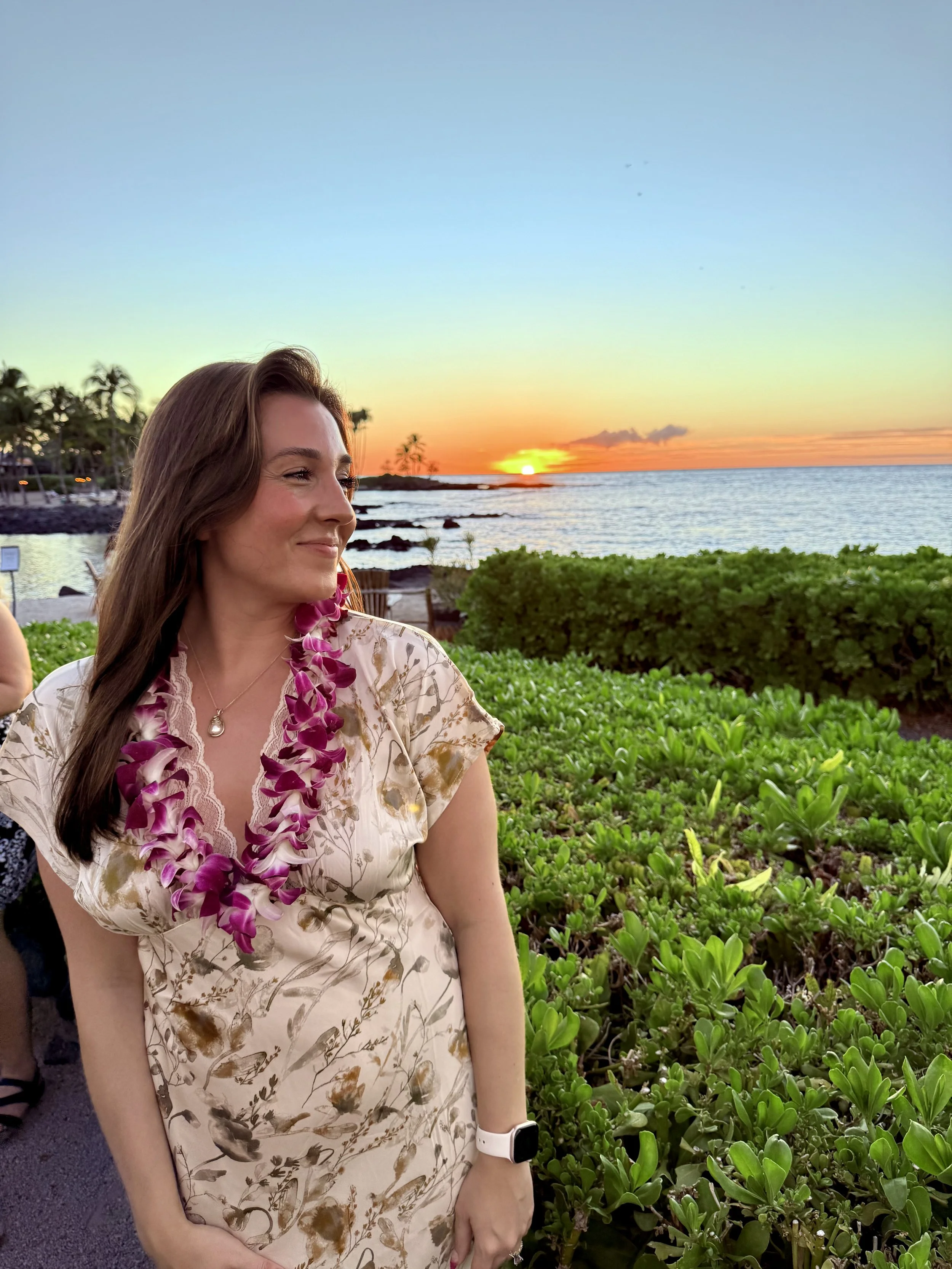 A woman wearing a floral dress and a purple lei stands outdoors during sunset near the ocean, surrounded by green foliage.
