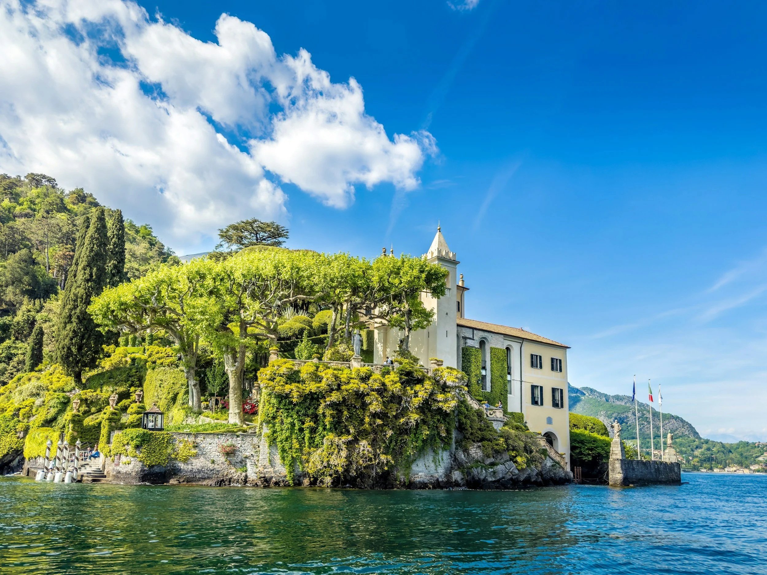 A scenic view of a villa on a lakeshore with lush green trees, a stone wall, and flags, under a partly cloudy blue sky.