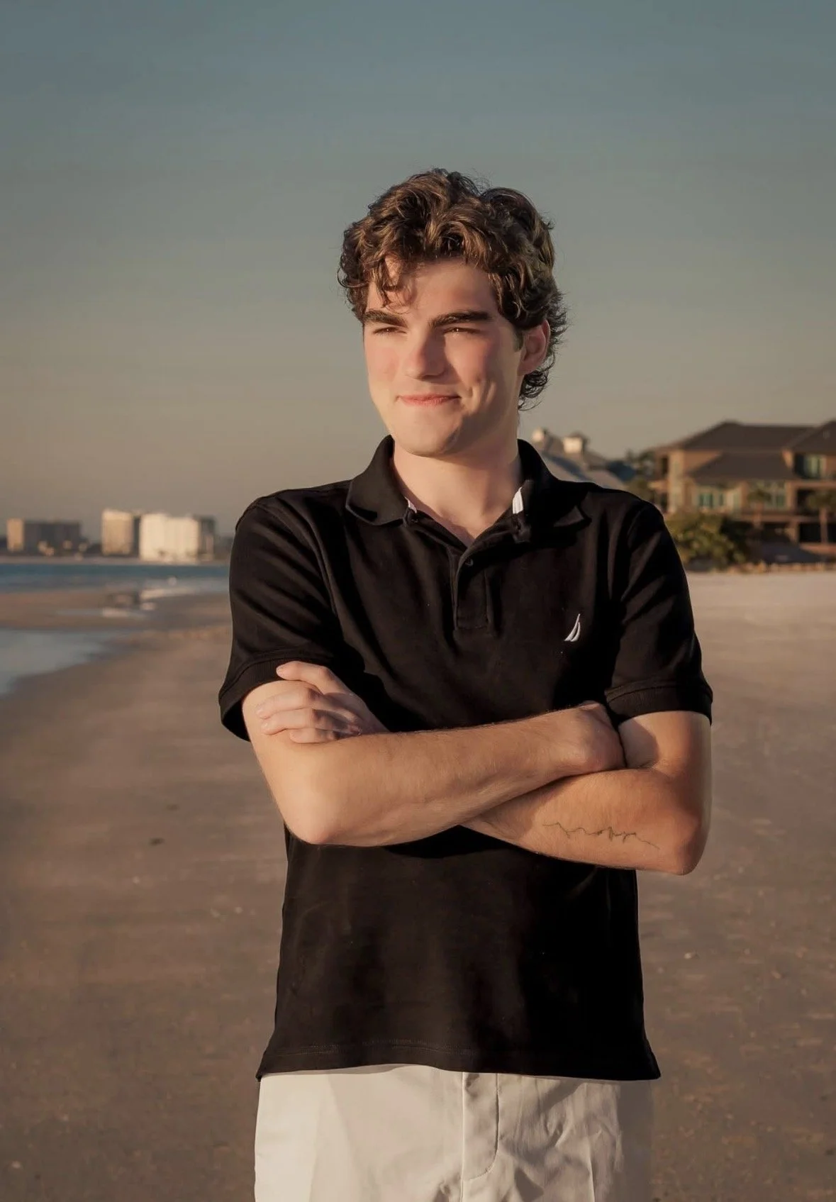 Young man standing on the beach with arms crossed, smiling, during sunset, with buildings in the background.