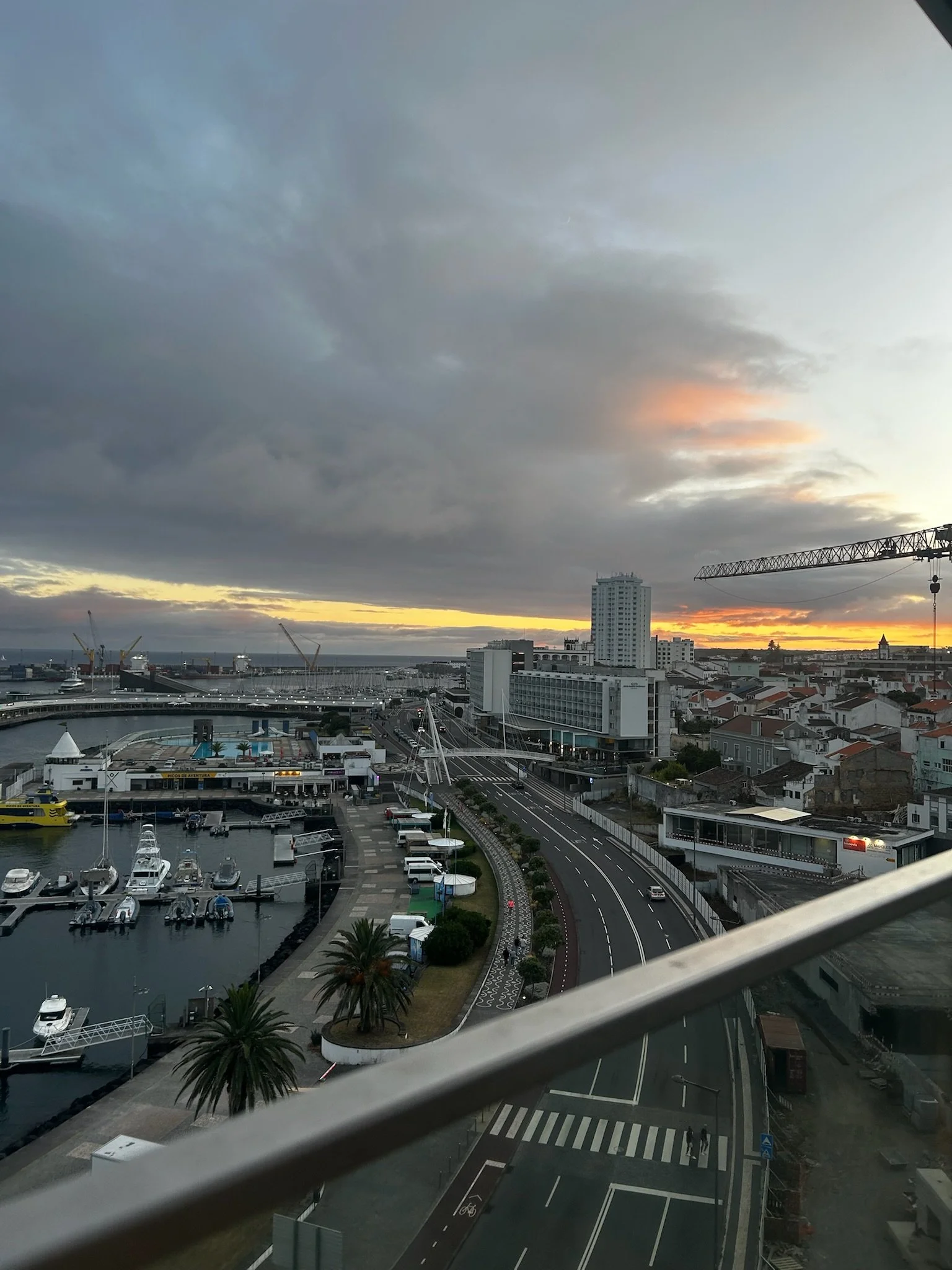 View of a cityscape at sunset with a harbor in the foreground, boats docked, high-rise buildings, and cloudy sky with a hint of orange glow.