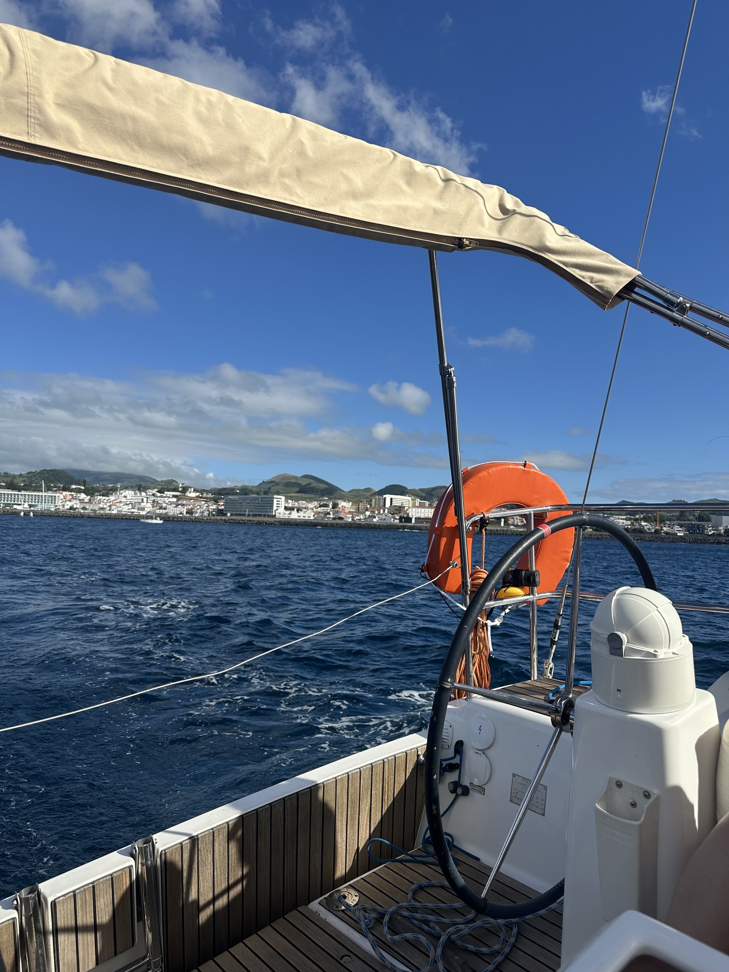 View from a sailboat showing a blue ocean, a distant shoreline with buildings, and a sky with a few clouds. The boat has a beige sail, orange life ring, and various ropes and equipment.