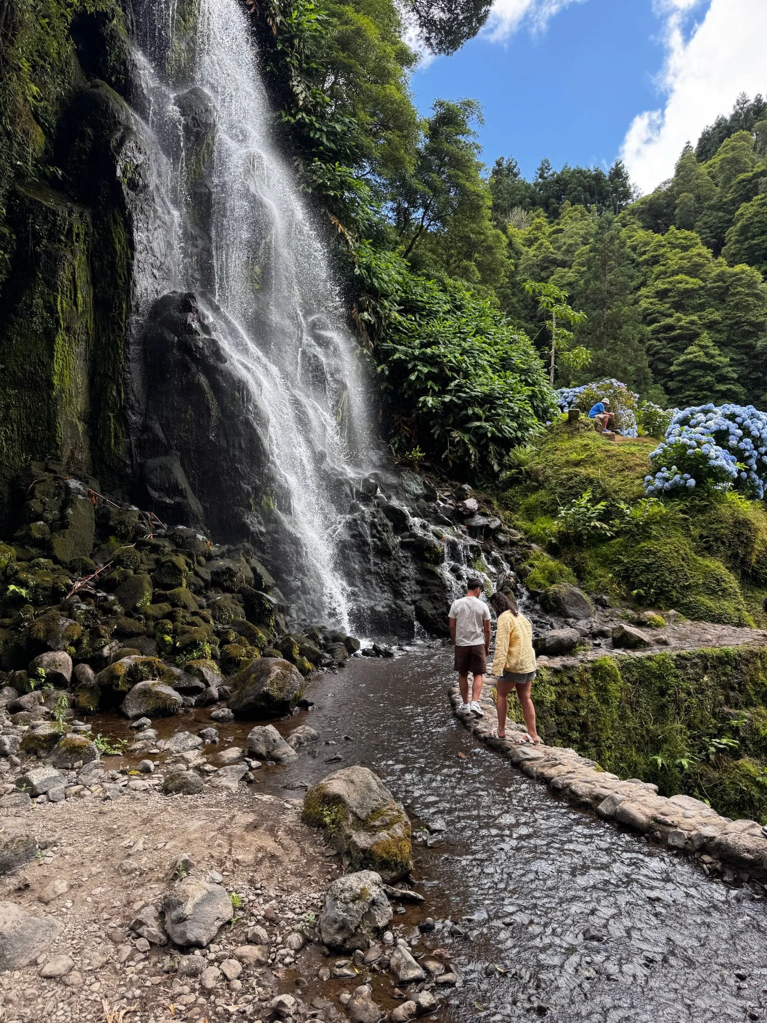 A waterfall cascading down a rocky cliff surrounded by green trees in a forest. Two people stand on rocks near a small stream at the bottom of the waterfall, looking at the scene, with another person sitting on a rock overlooking the waterfall.