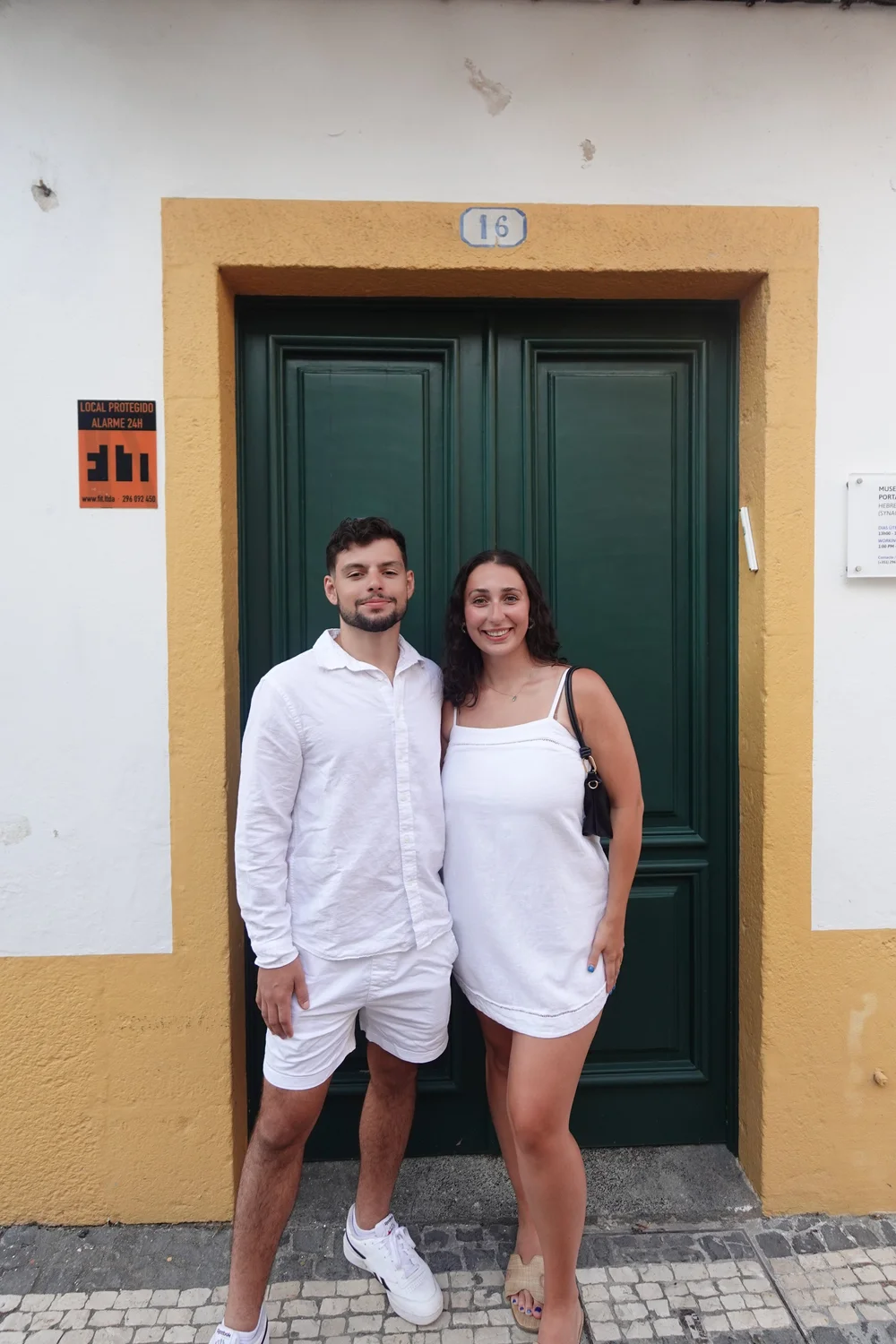 Mateus and his partner smiling in front of a green door with a yellow frame on a cobblestone street in São Miguel, Azores.