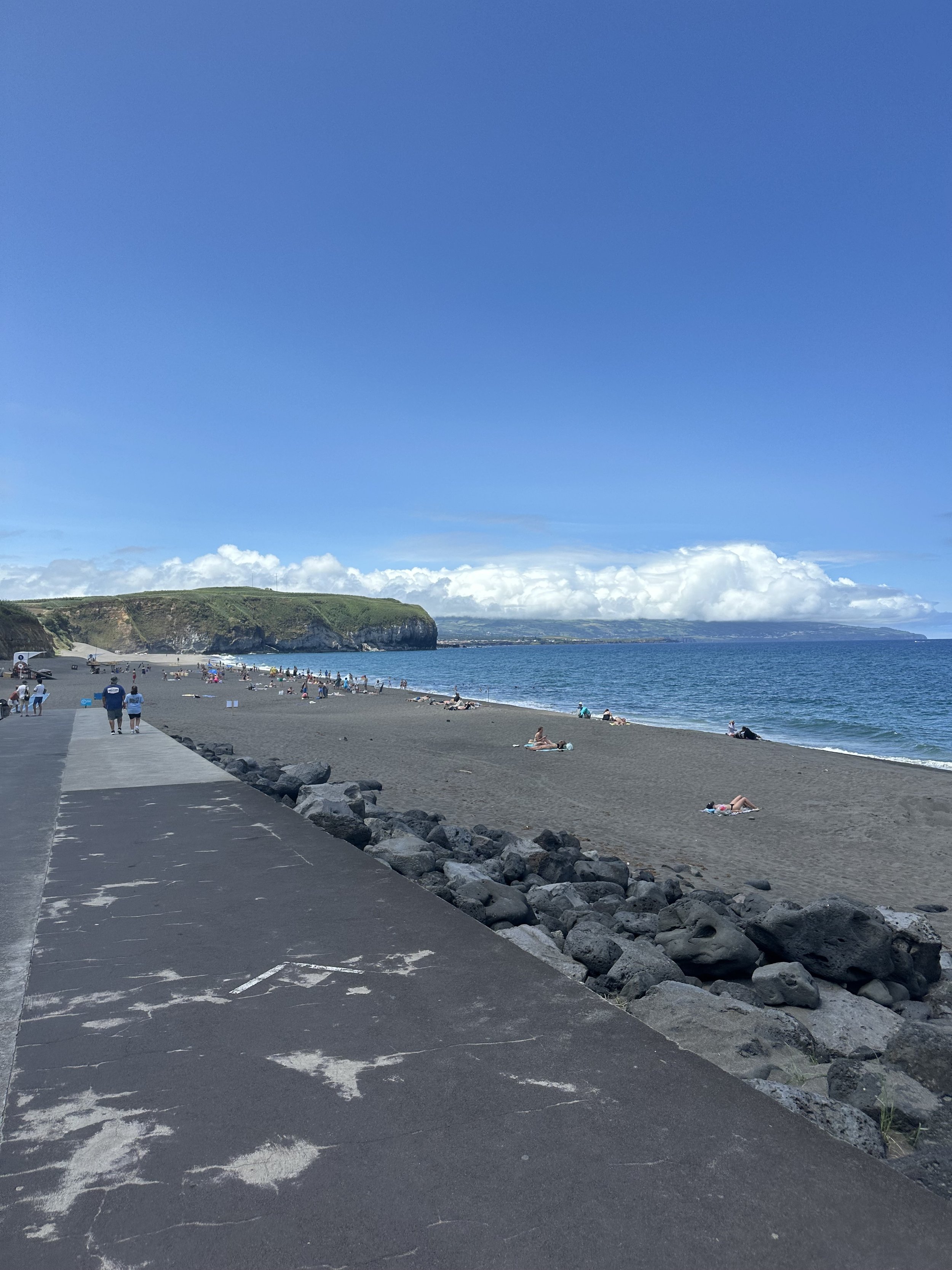 A beach with dark sand, people relaxing and walking, a rocky pathway, a cliff with greenery, and a blue sky with clouds over the ocean.